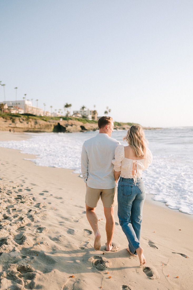 Couple walking barefoot together along sandy shoreline with palm trees dotting the coastal cliffs.