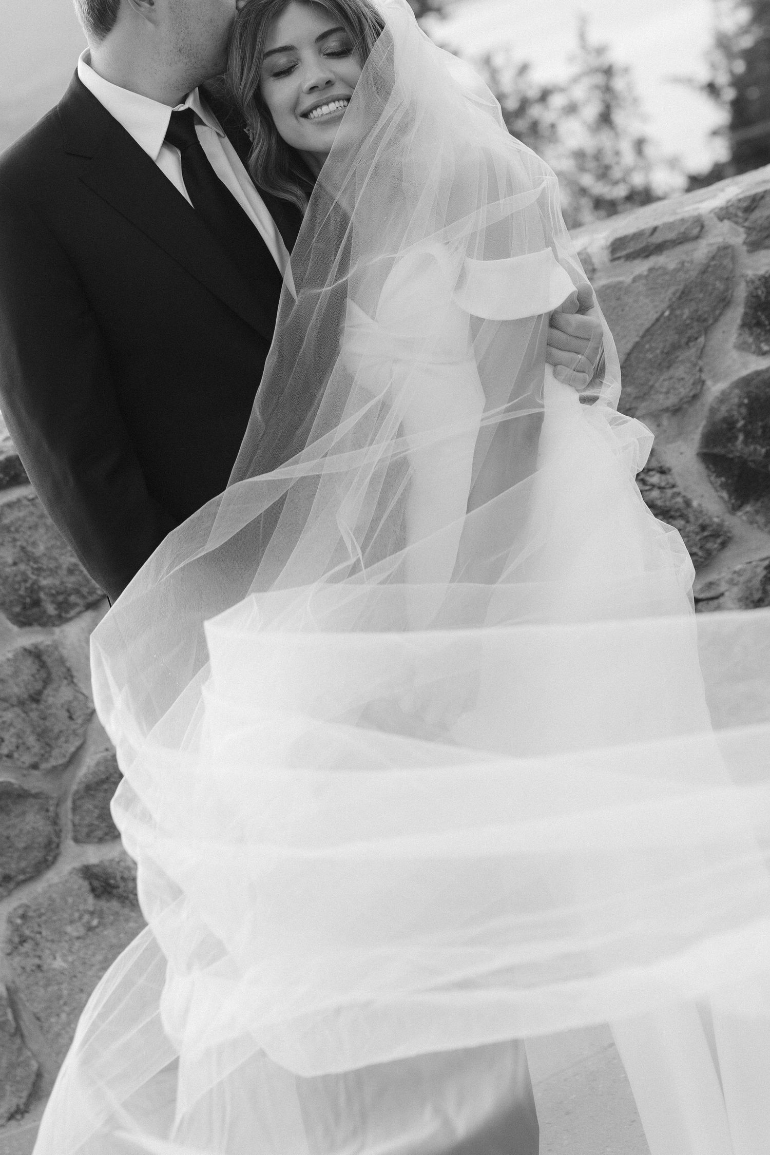 A black and white wedding photograph showing a flowing white veil and dress against a stone wall.