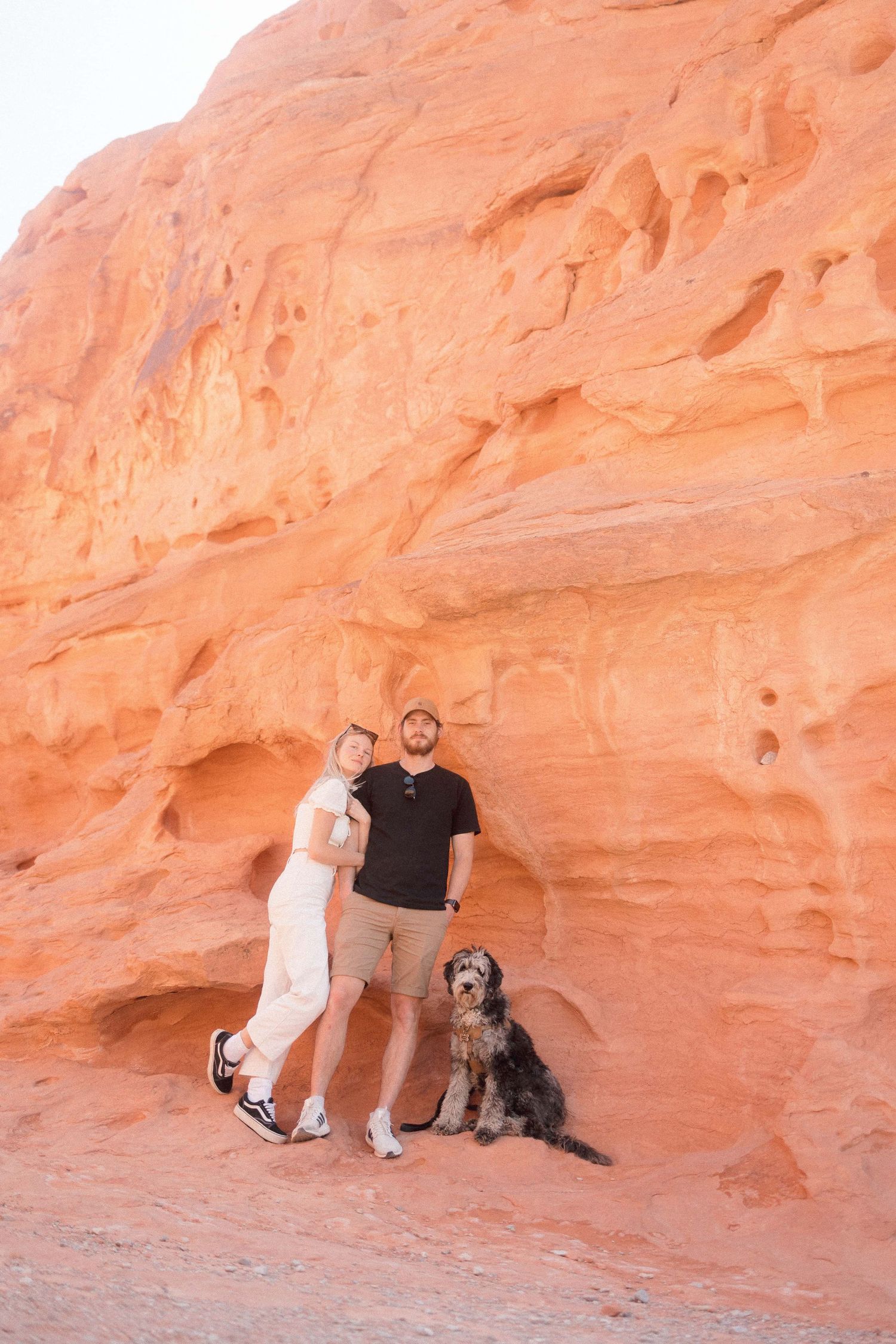 A dog sits with two people against a towering orange sandstone wall in a desert landscape.
