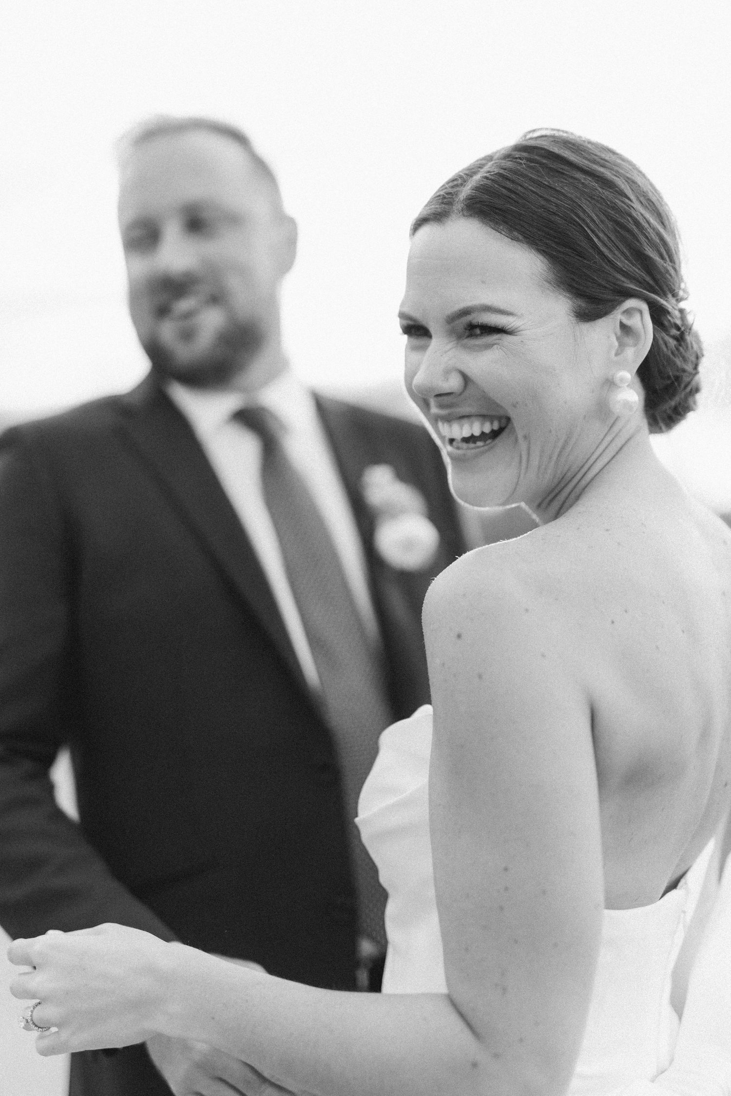 Black and white candid wedding photo showing a joyful bride laughing while groom stands in background.