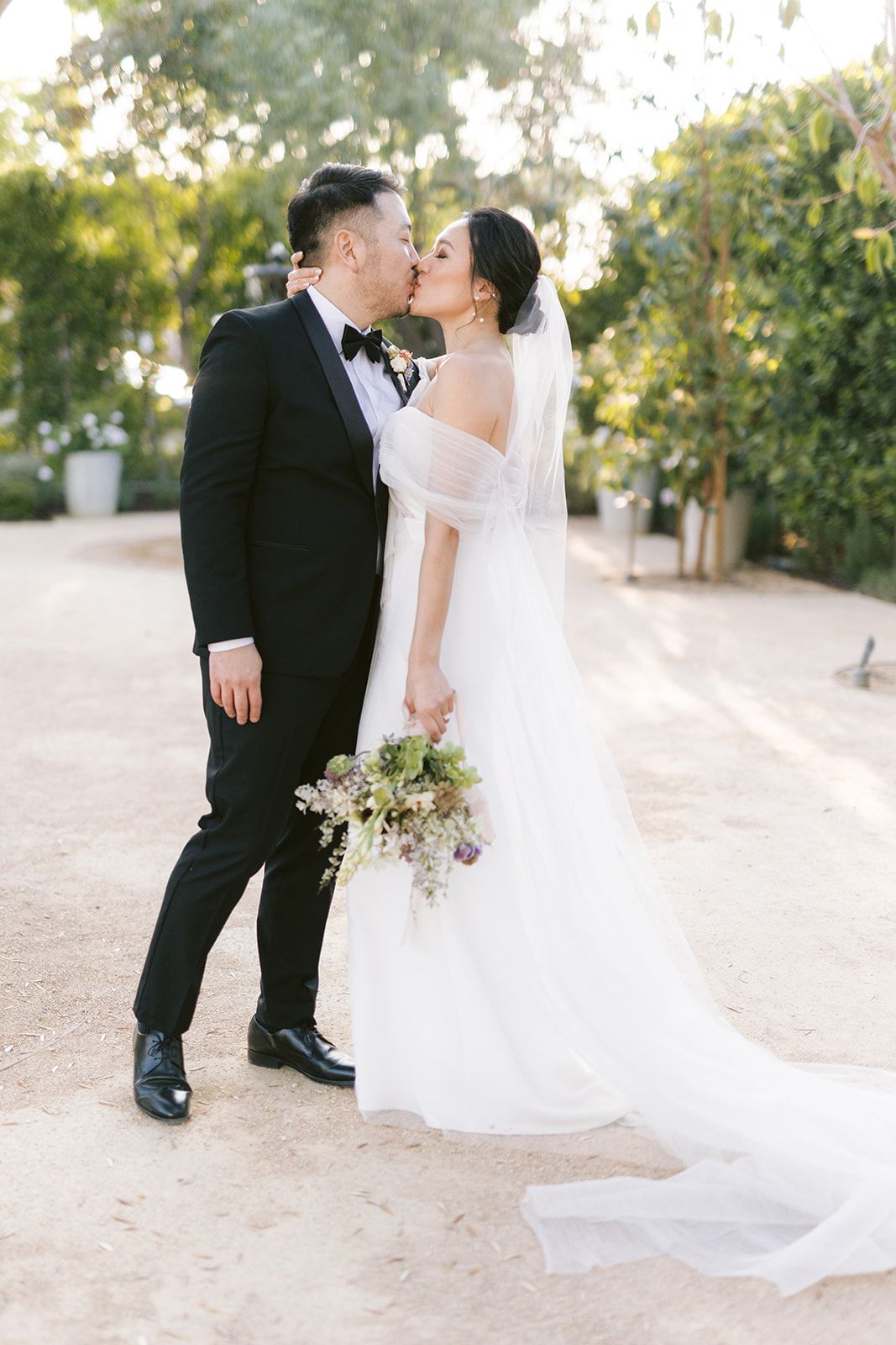 A romantic wedding couple shares a tender kiss on a sunlit garden path while the bride holds a delicate bouquet.