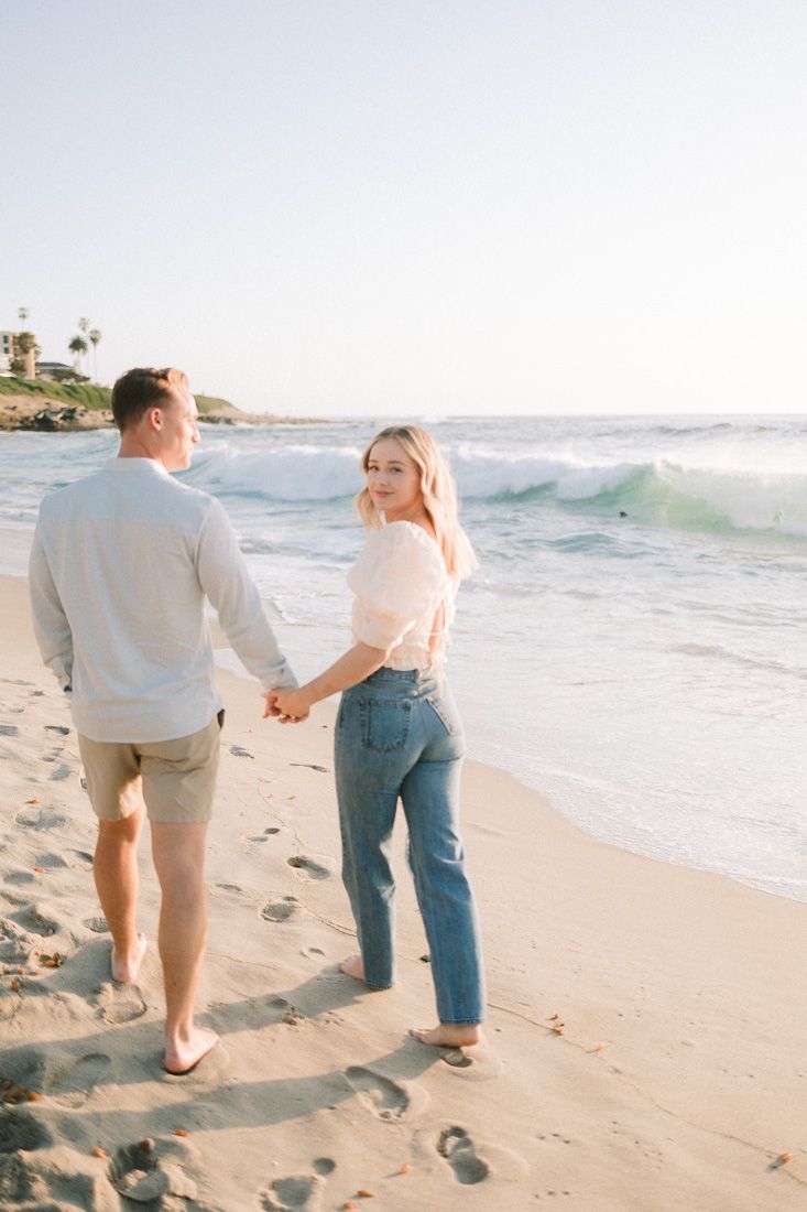 Romantic beach stroll with couple holding hands against crashing waves and coastal cliffs.