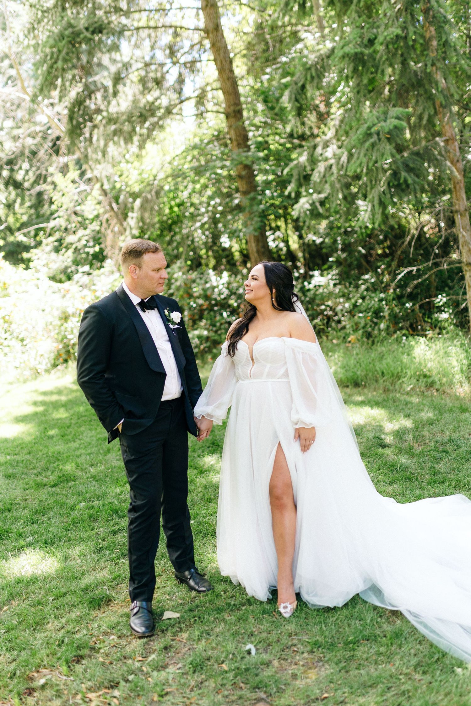 A couple in wedding attire stands together on grass with evergreen trees in the background.