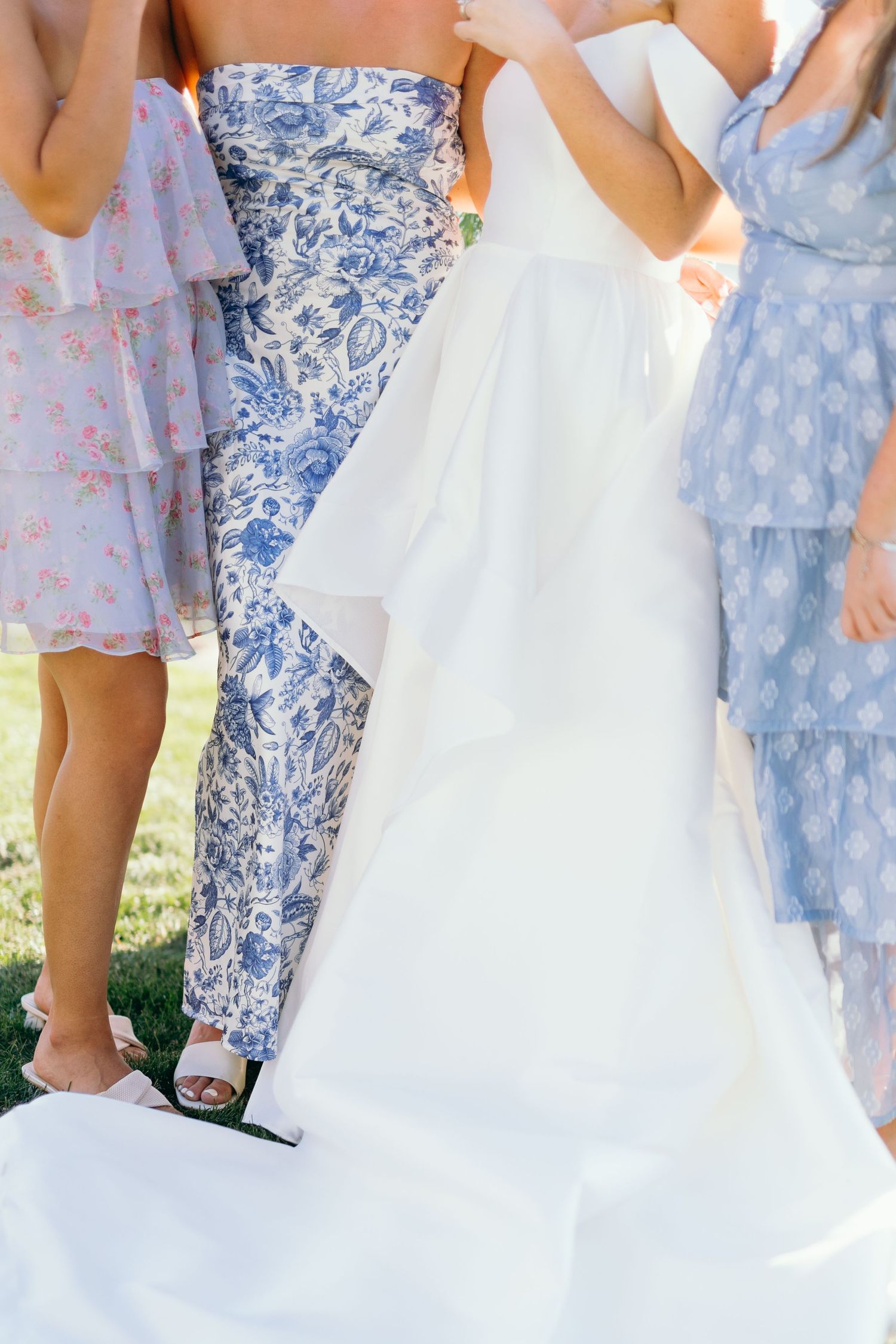 Bridesmaids in floral blue and pink dresses stand beside a flowing white wedding gown outdoors.