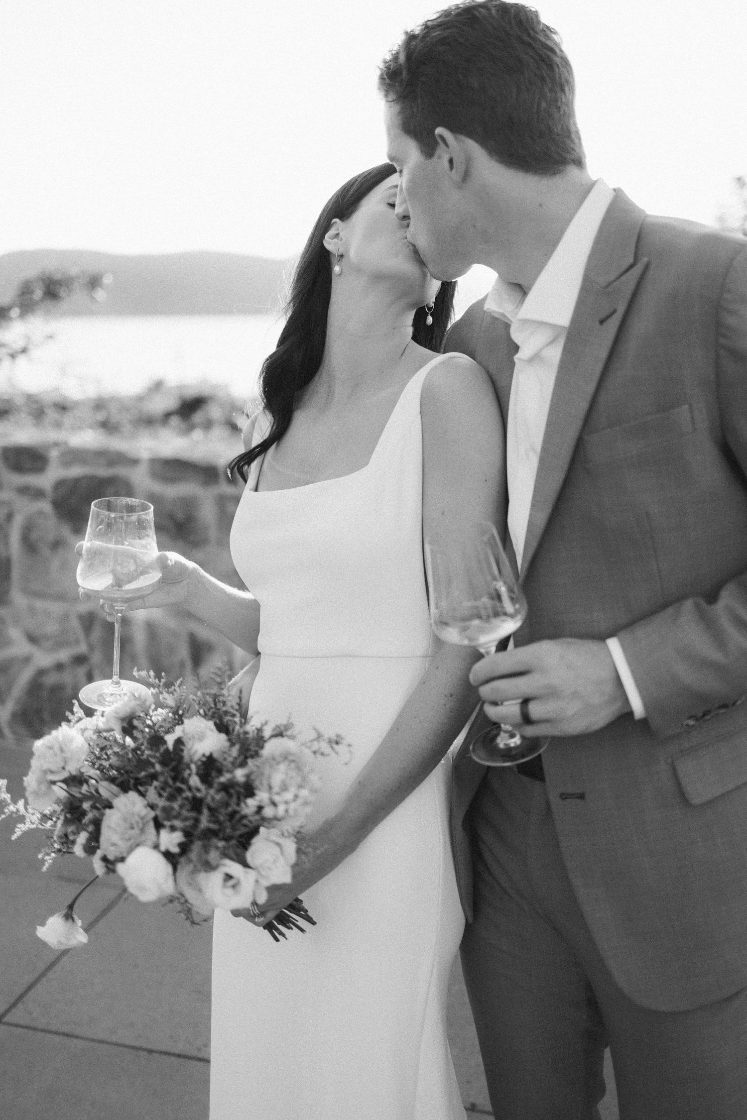Black and white wedding photo of couple sharing a kiss while holding champagne glasses and bouquet by the water.