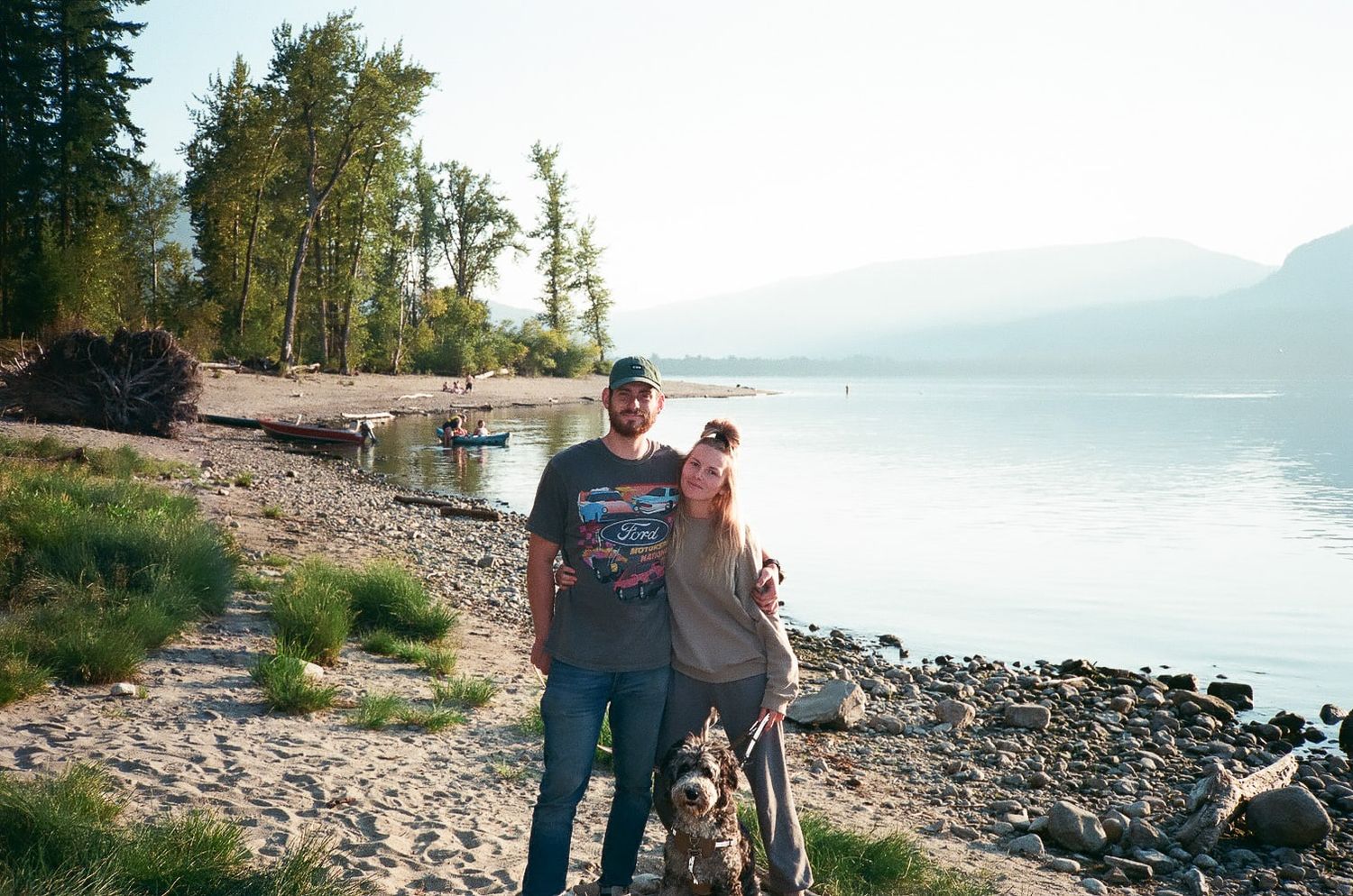 Two people stand together on a rocky beach shore with mountains and a serene lake in the background.