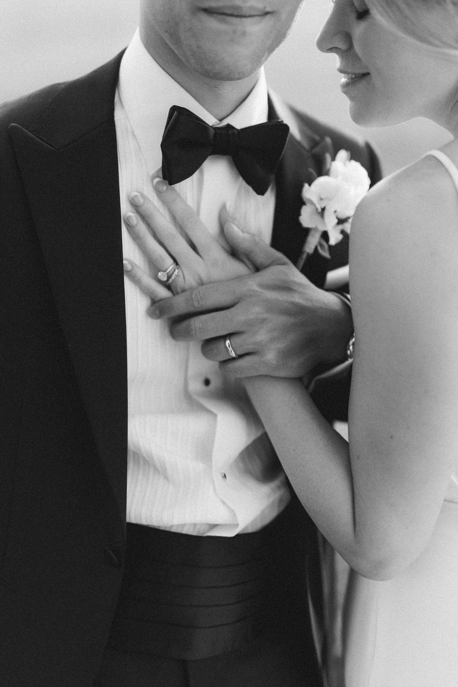 Black and white close-up of newlyweds showing their wedding rings with the groom in a tuxedo and white boutonniere.
