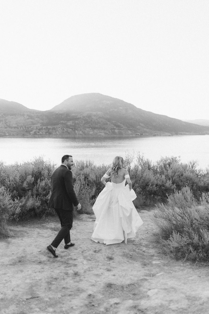 A couple in wedding attire walks along a dirt path near a serene lake surrounded by mountains in black and white.