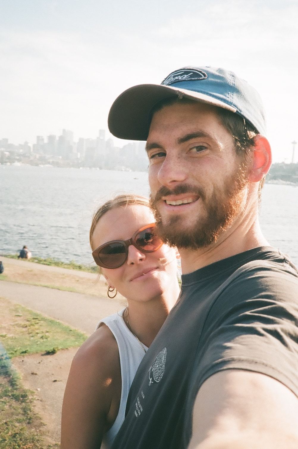 A couple takes a waterfront selfie on a sunny day with a city skyline visible in the background.