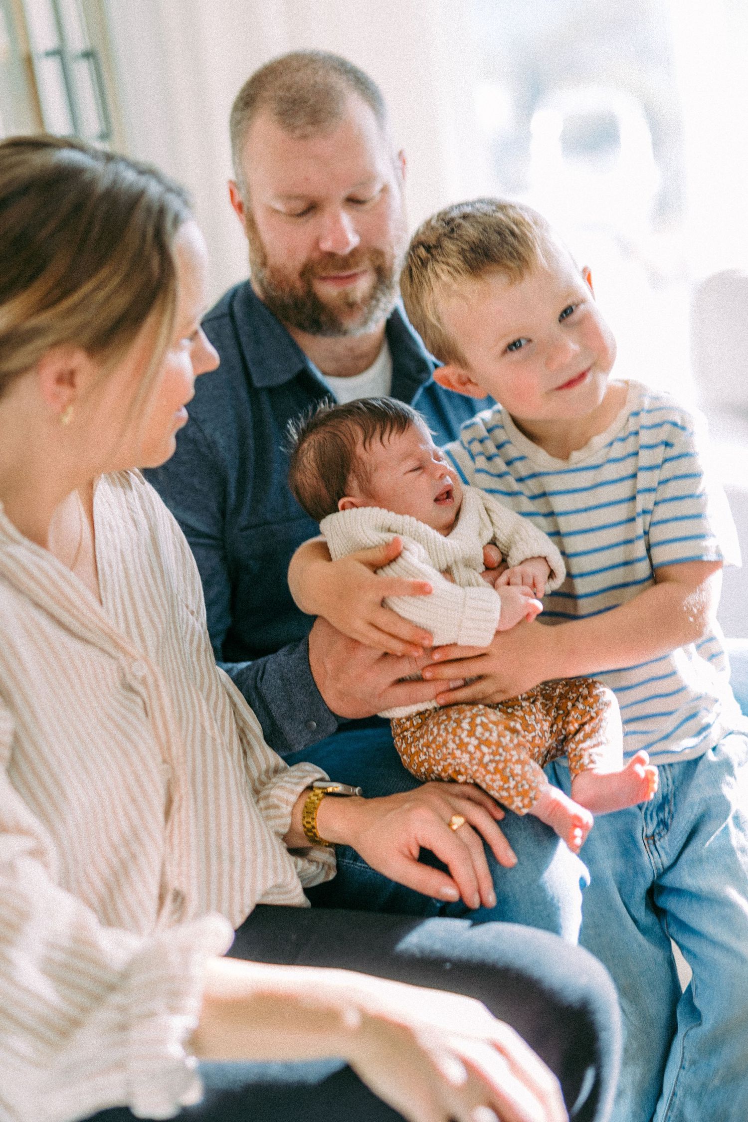A family sits together on a couch, sharing a tender moment with their newborn baby and young child in a bright room.