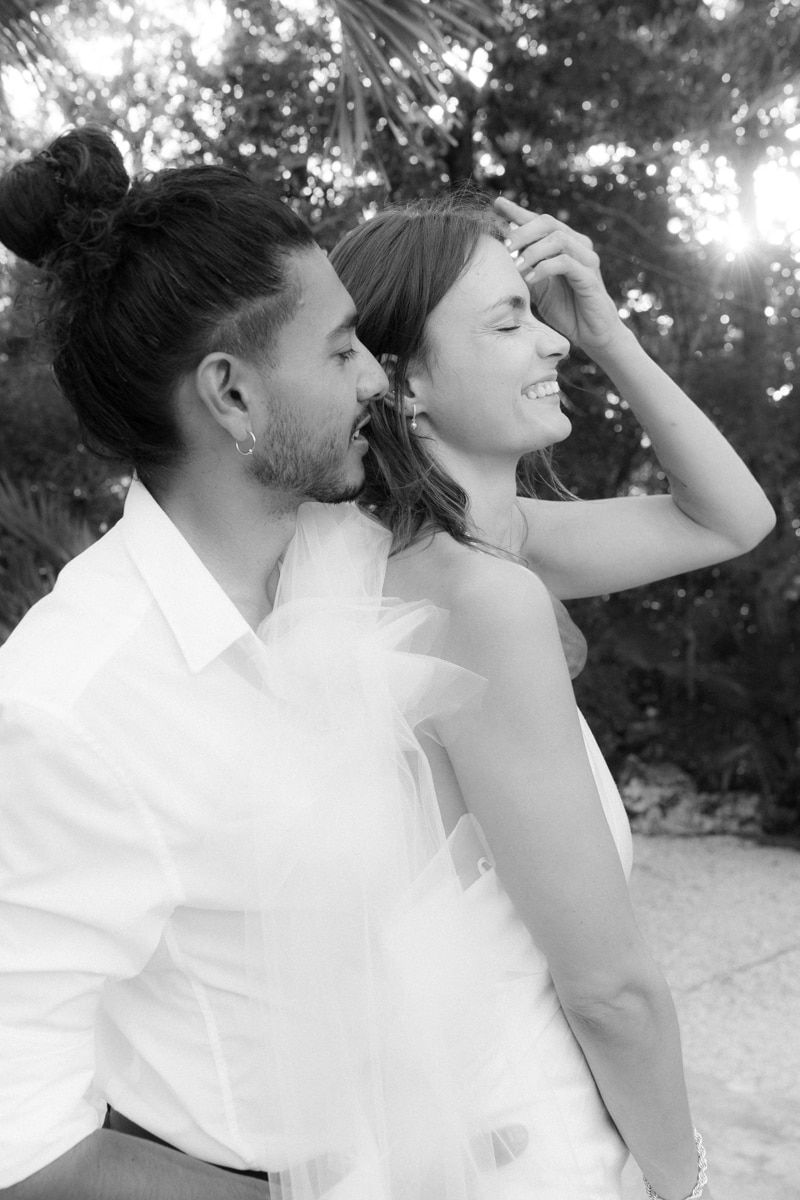 A romantic black and white portrait of a couple sharing a joyful moment outdoors with sunlight filtering through trees.