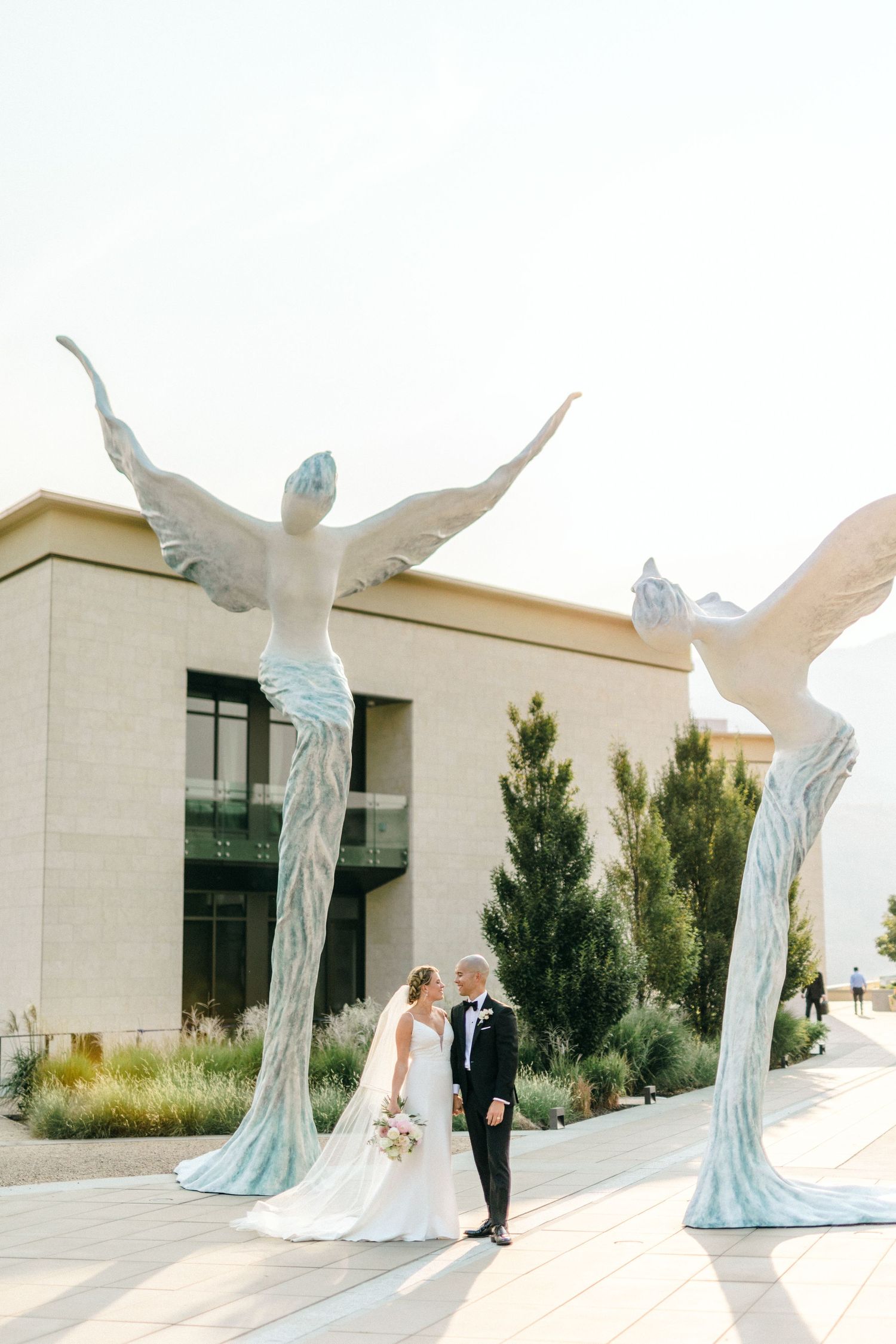 A couple poses beneath large sculptural angel wings in pale blue and white outside a modern building.