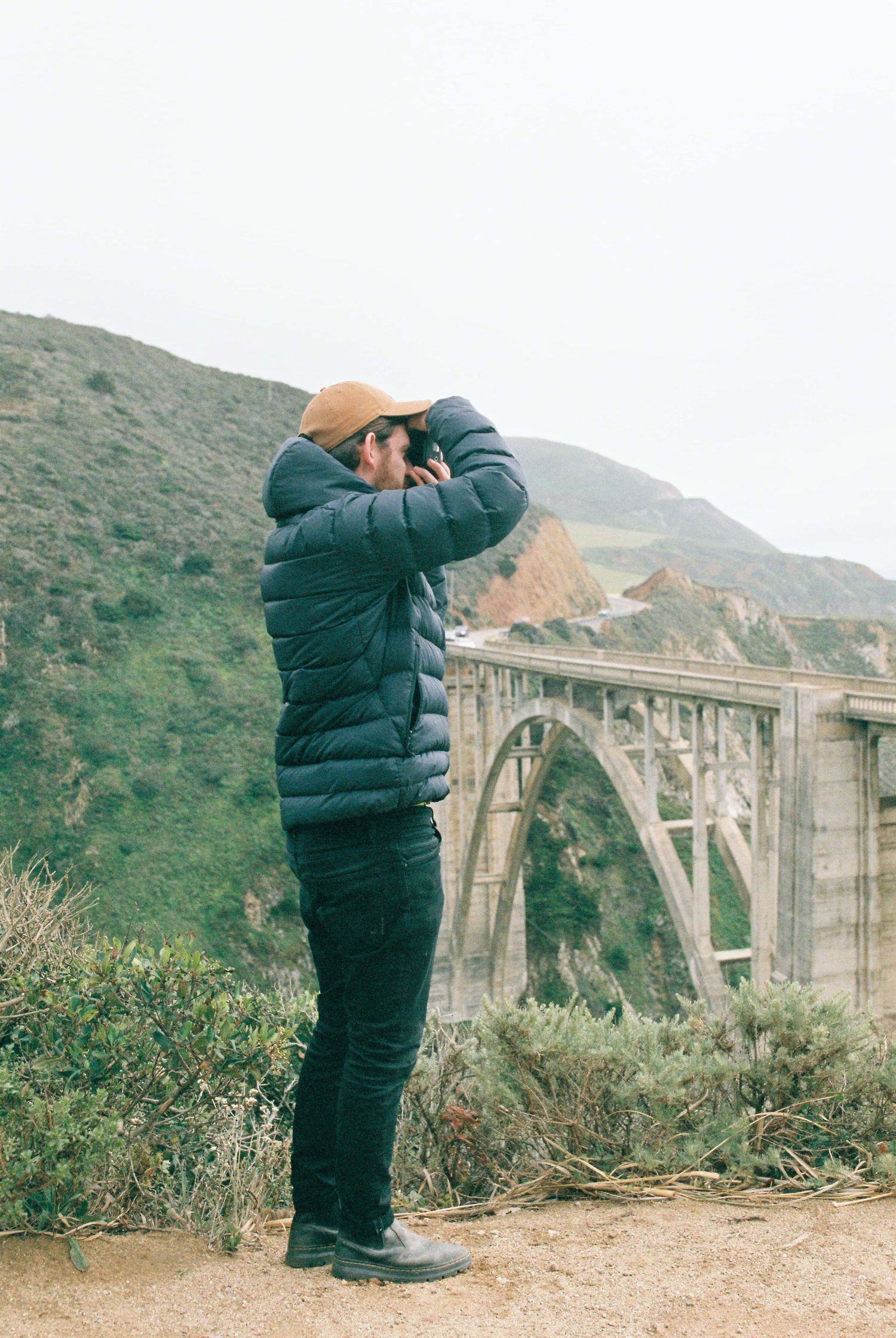 Person in a blue puffer jacket takes photos at Bixby Creek Bridge in Big Sur, California with coastal mountains in view.