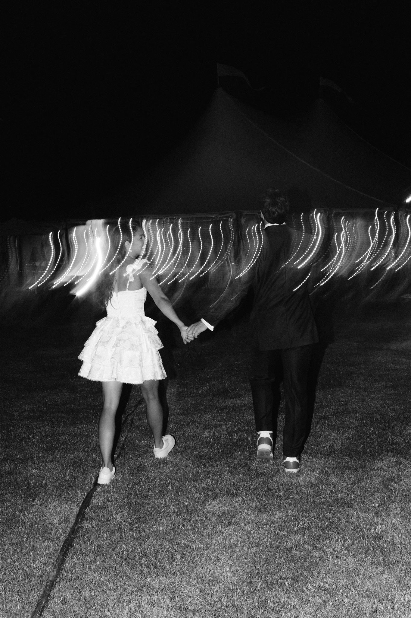 A black and white long exposure photo shows streaks of light above a couple walking on grass at night.