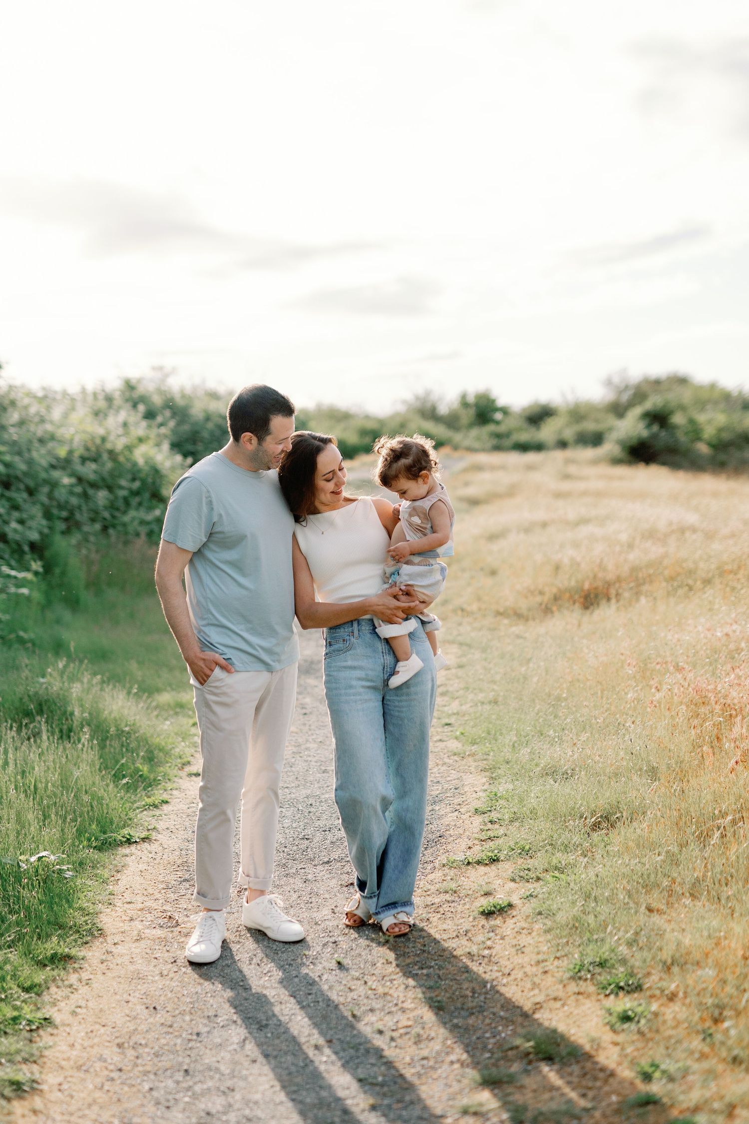 A family walking together on a dirt path through a sunny meadow wearing casual clothes on a summer day.