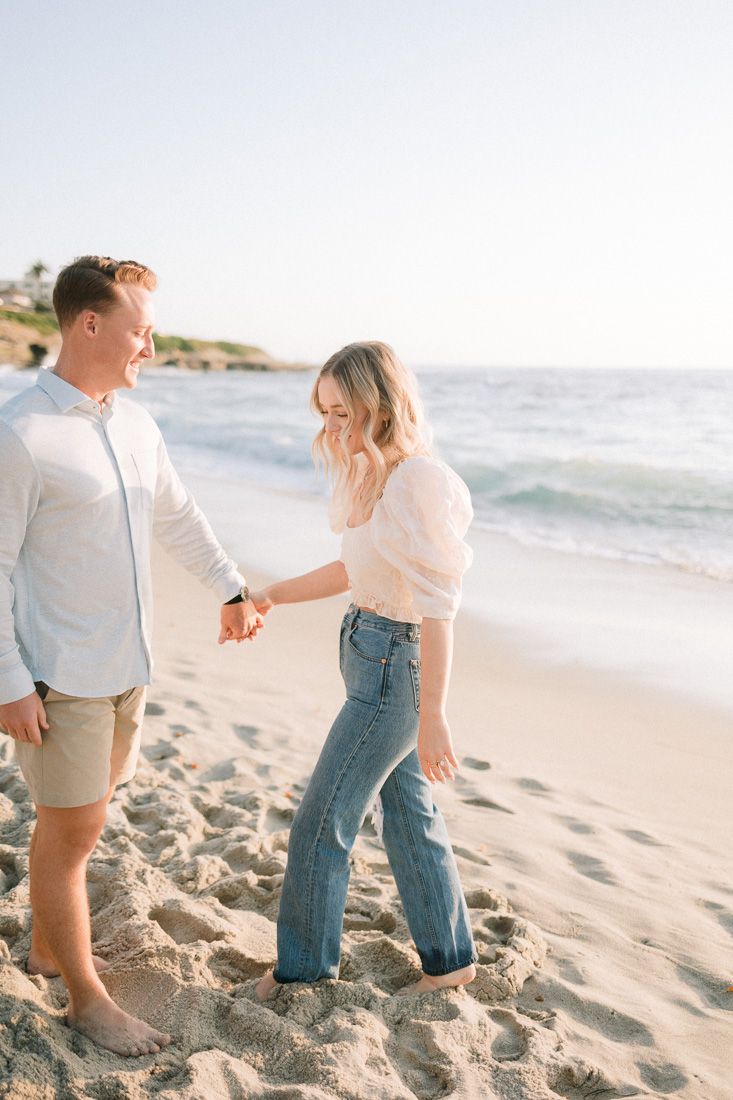 Couple sharing playful moment on sandy beach with ocean waves and cliffs in background.