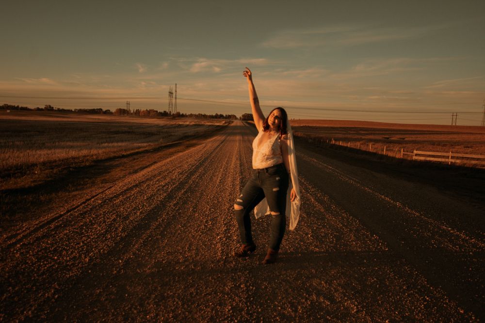 A silhouetted figure celebrates with arms raised on a rural dirt road at sunset.