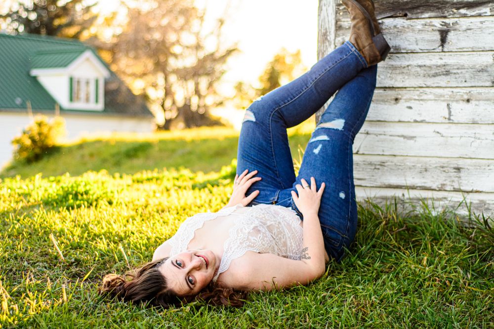A white lace-clad figure lying in green grass next to a rustic wooden wall at sunset.