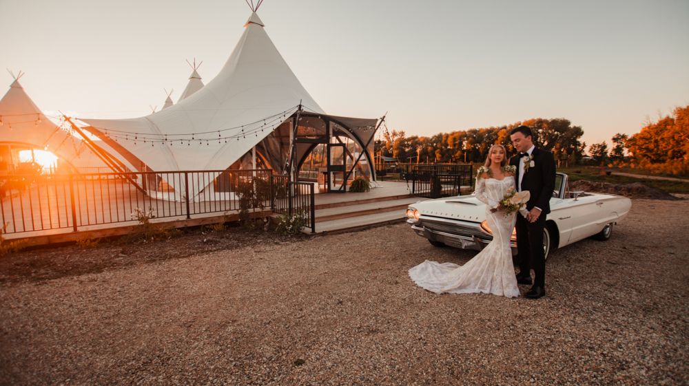 Wedding couple poses outside reception tent at sunset with classic white convertible car.