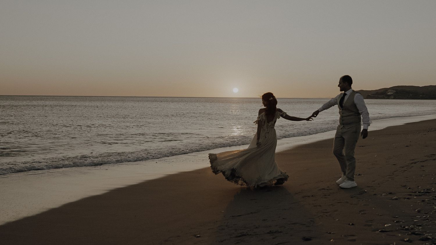 Silhouetted couple holding hands on beach at sunset during wedding.