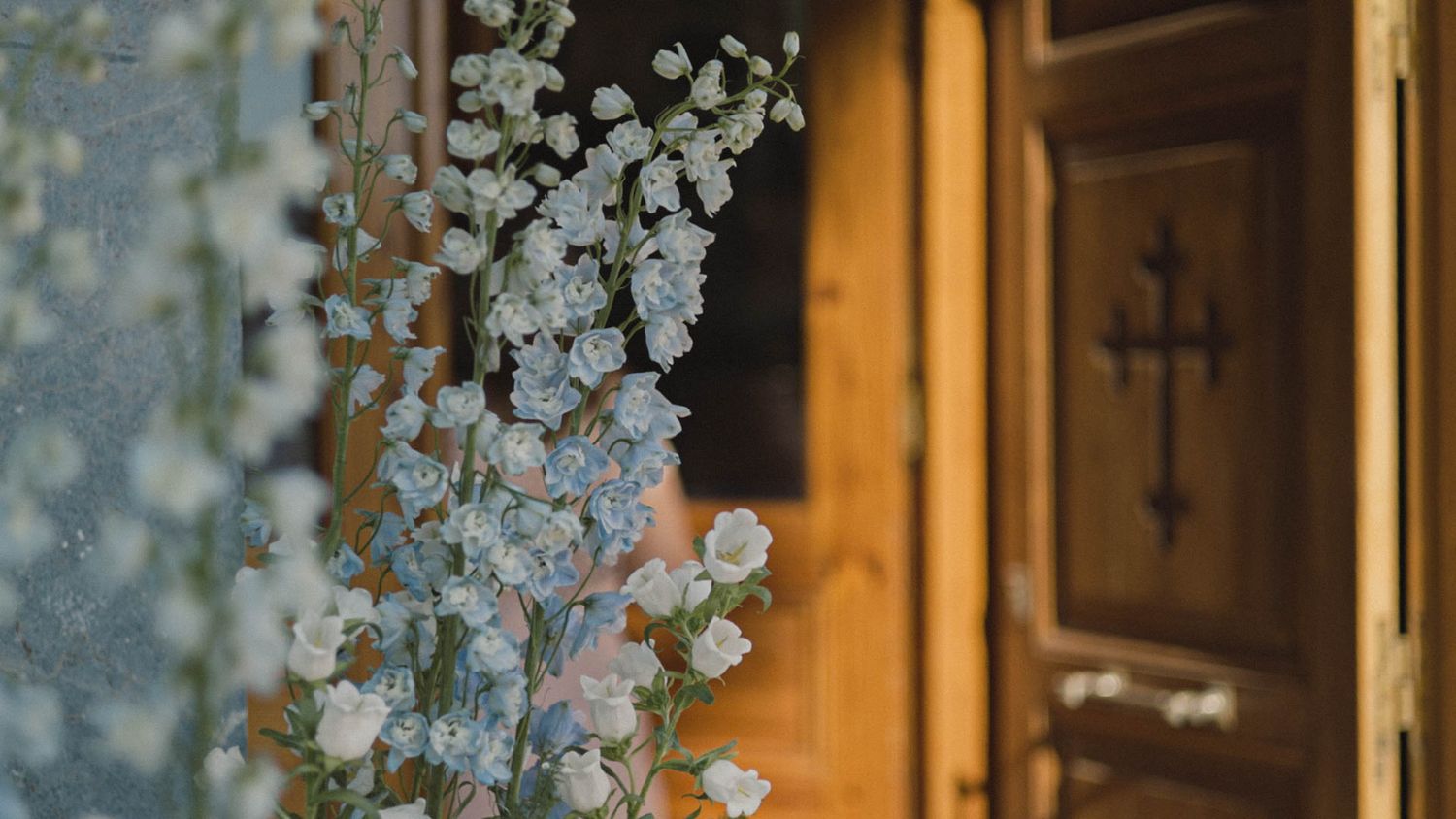 Delicate white flowers bloom near a wooden door with a cross design in warm lighting.