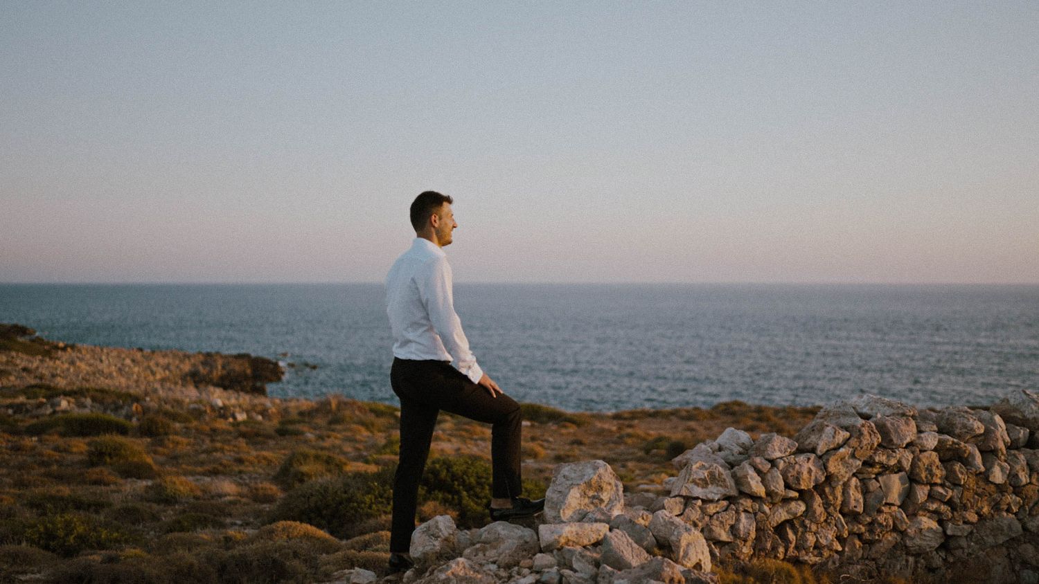 A figure in a white sweater stands on rocky coastal cliffs overlooking a calm ocean at dusk.