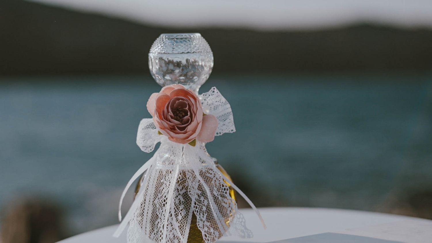 Small decorative glass bottle adorned with delicate lace and a peach-colored rose against a blurred waterfront backdrop.