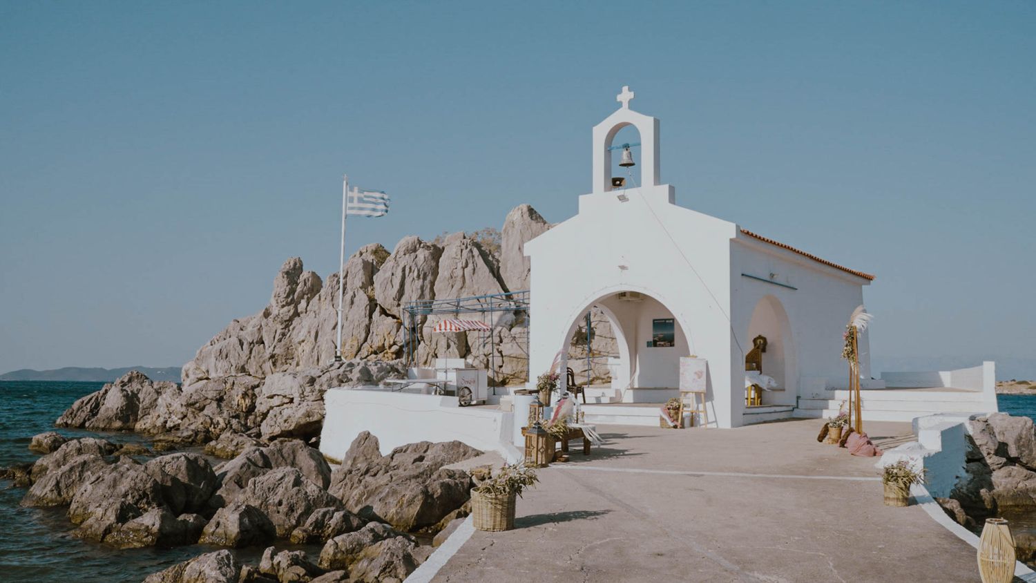 A small white Greek Orthodox chapel with a bell tower perches on rocky coastline overlooking the Mediterranean Sea.
