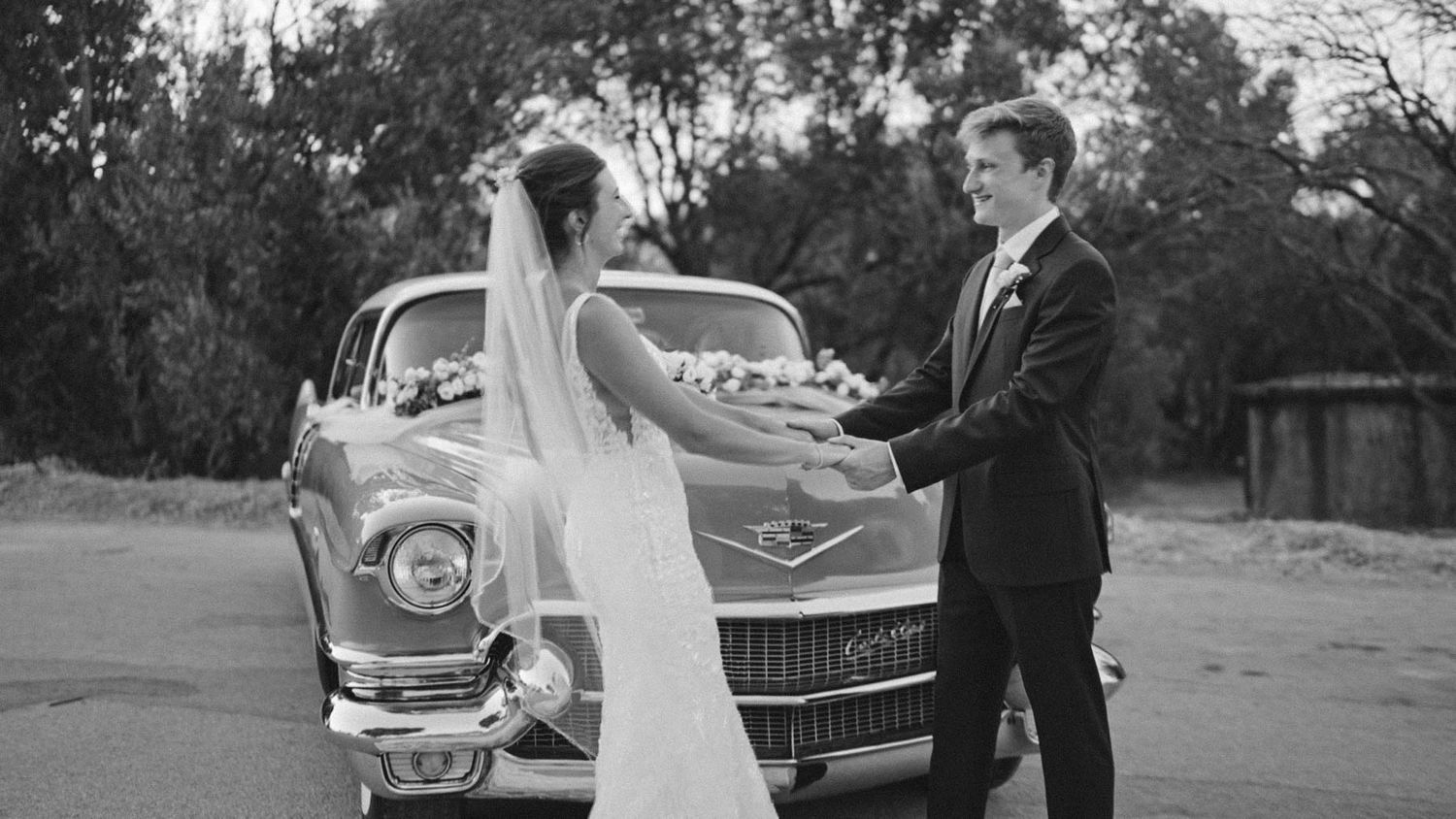 A classic 1950s vintage car serves as backdrop for a black and white wedding photograph.