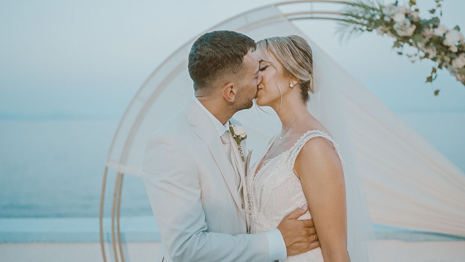 A happy couple shares intimate moments during their beach wedding ceremony against a pastel sunset backdrop.