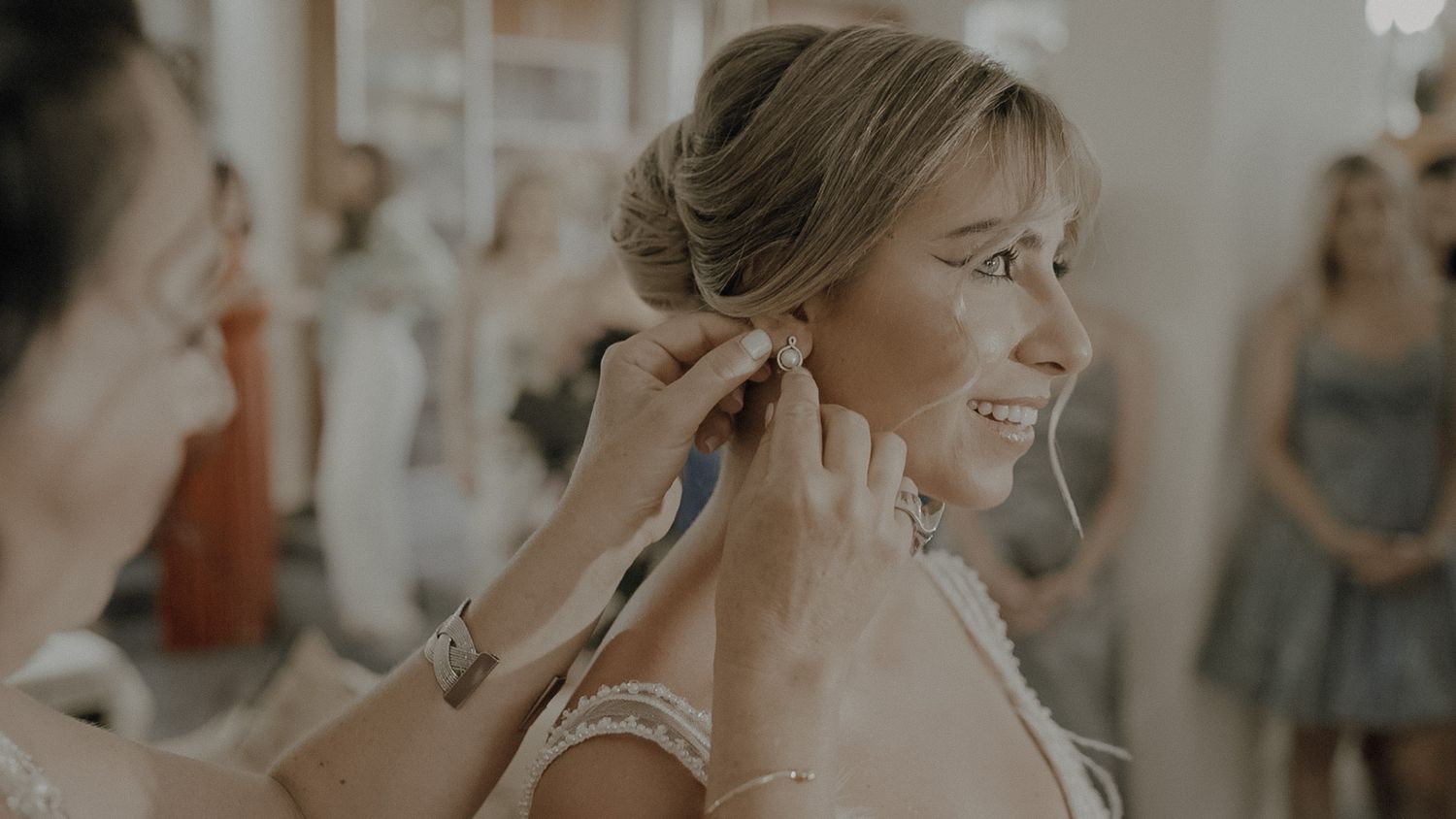Bride getting her earrings put on while smiling during wedding day preparations in warm lighting.