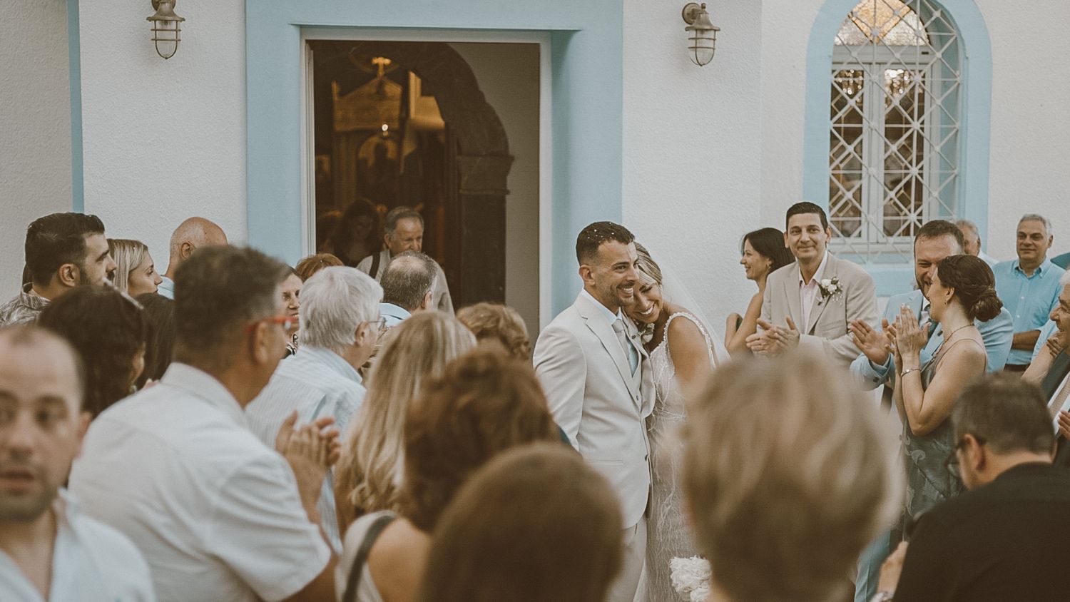A crowd of wedding guests gather outside a white church entrance during a joyful celebration.