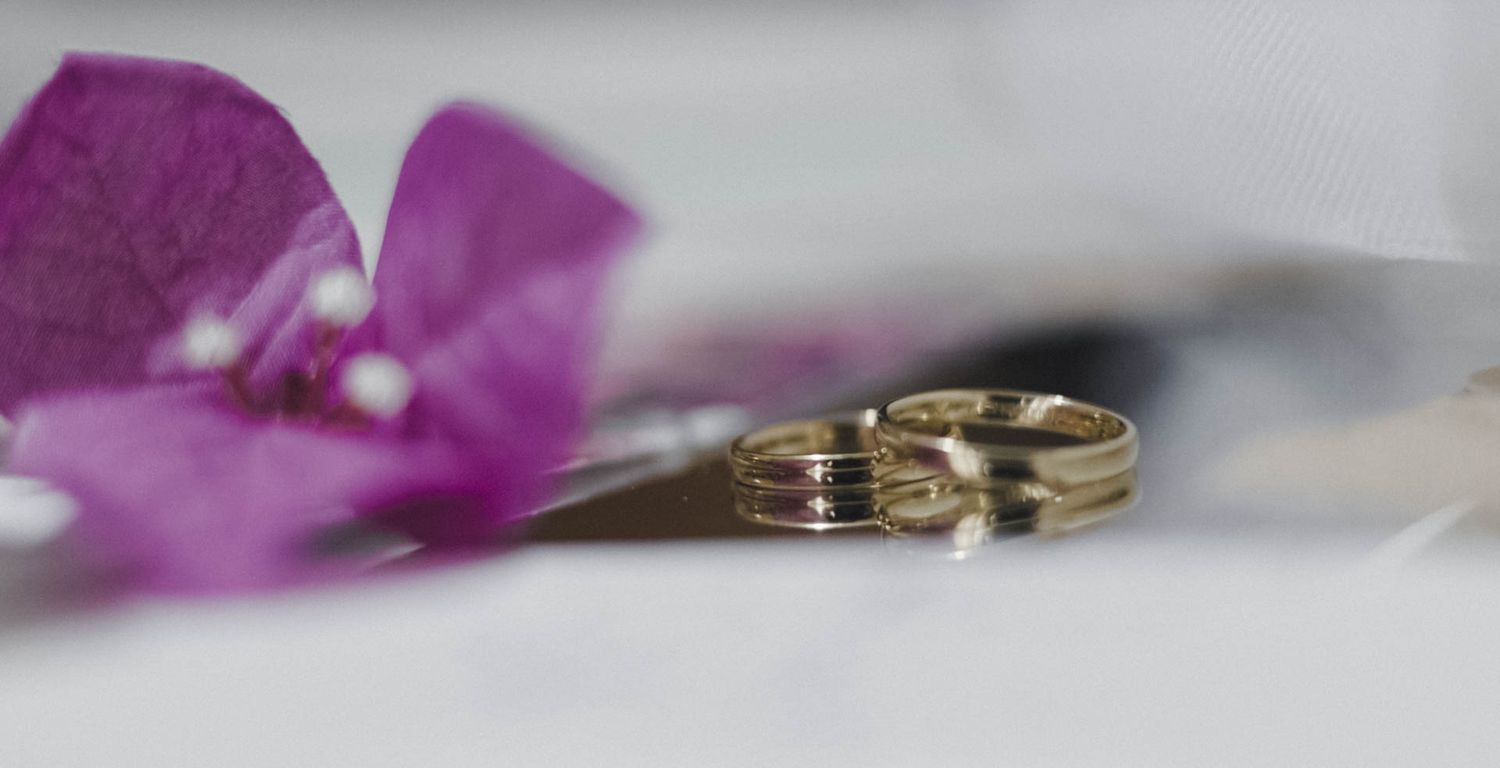 Two gold wedding rings displayed next to a purple orchid flower on a white surface in soft focus.
