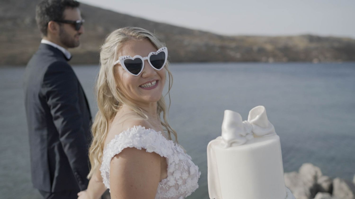 A bride in a white dress and sunglasses smiles near a three-tiered white wedding cake by the water.