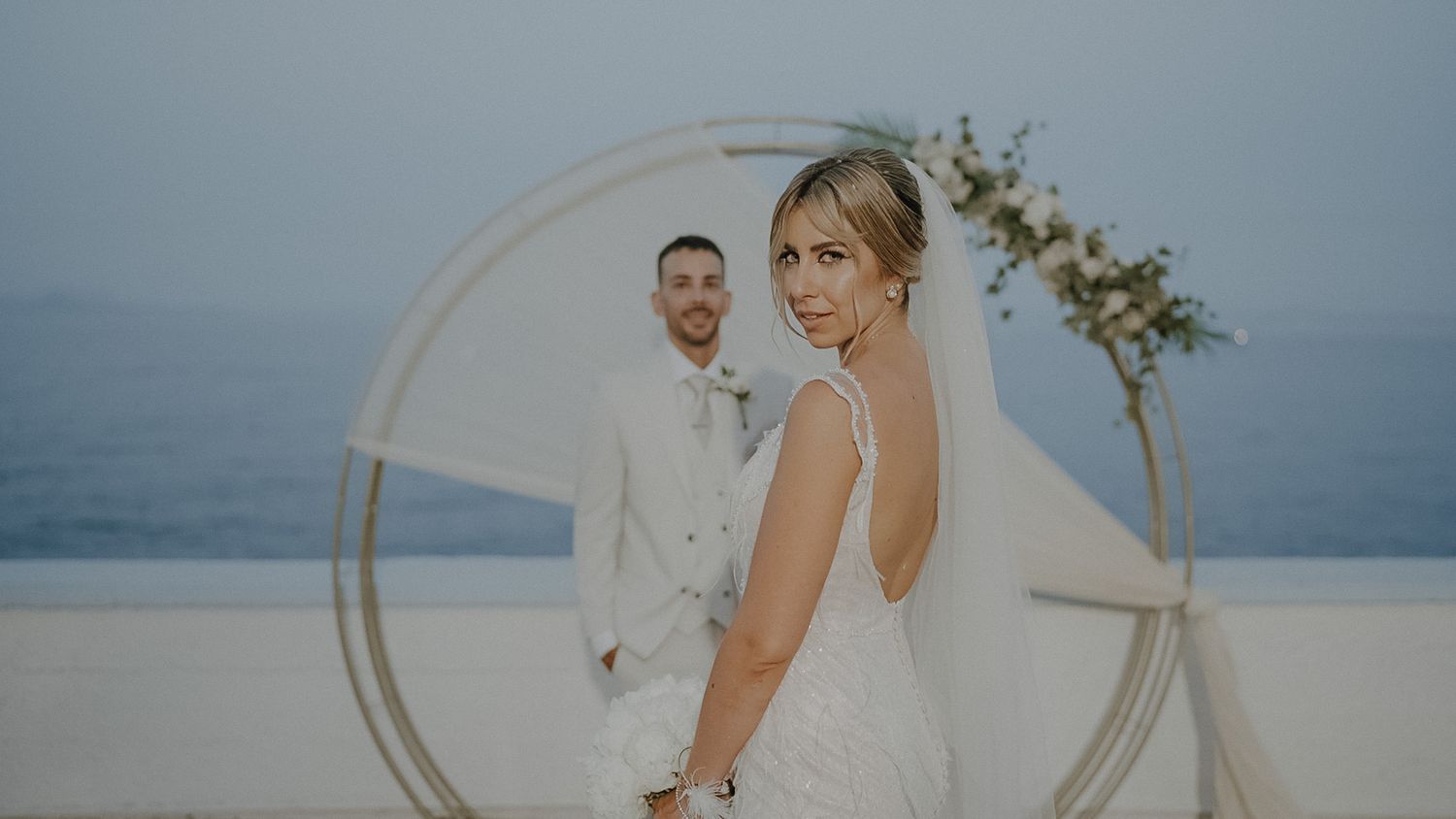 Elegant wedding celebration with white circular arch overlooking the ocean at sunset.