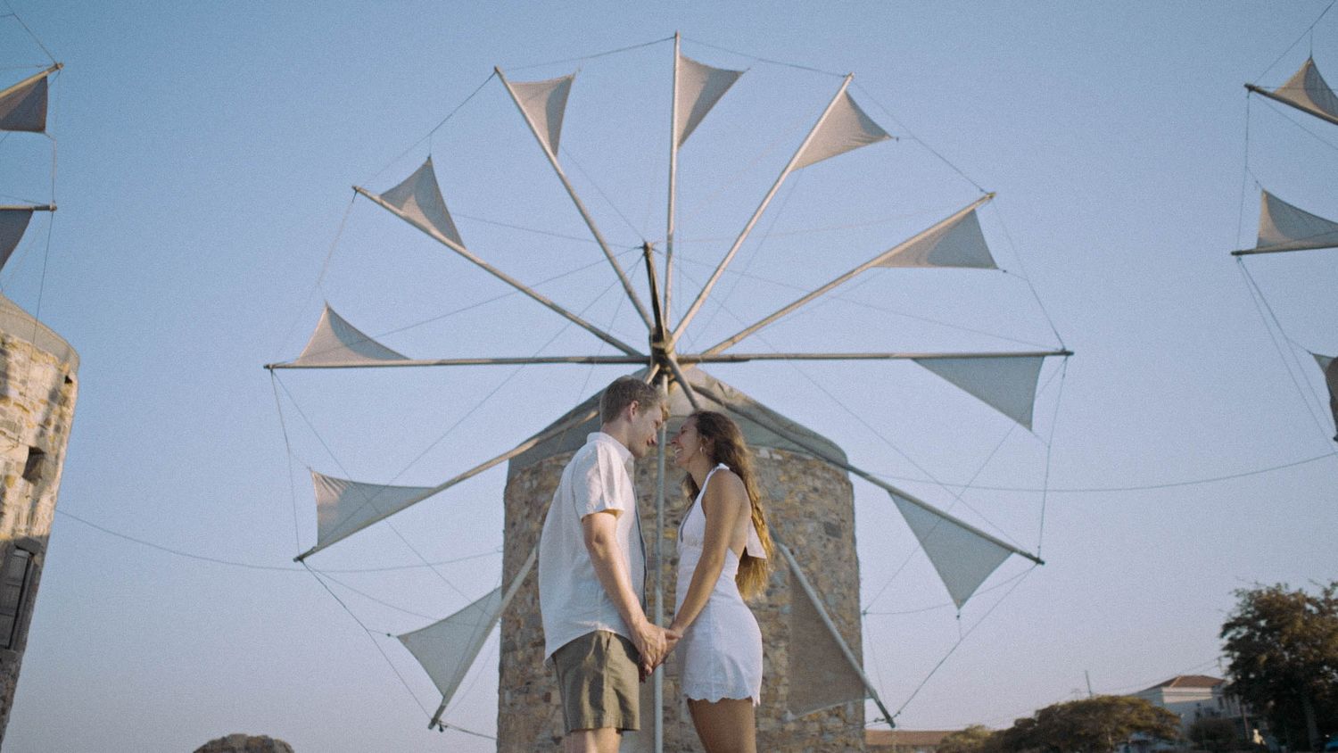A traditional Greek windmill with large white sail-like blades stands against a blue sky in Greece.