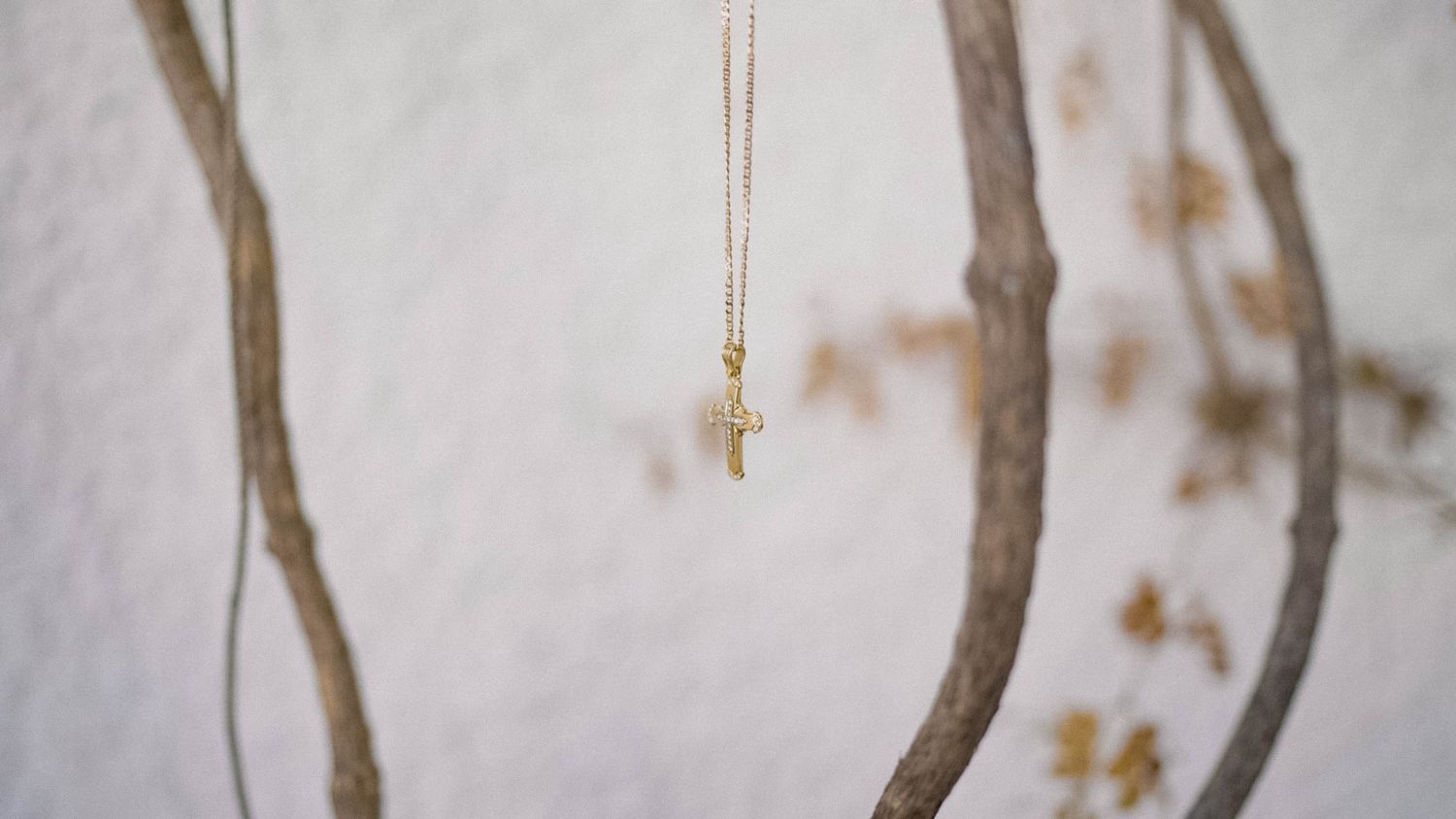 A small golden cross pendant necklace hangs delicately against a soft blurred background with curved branches.