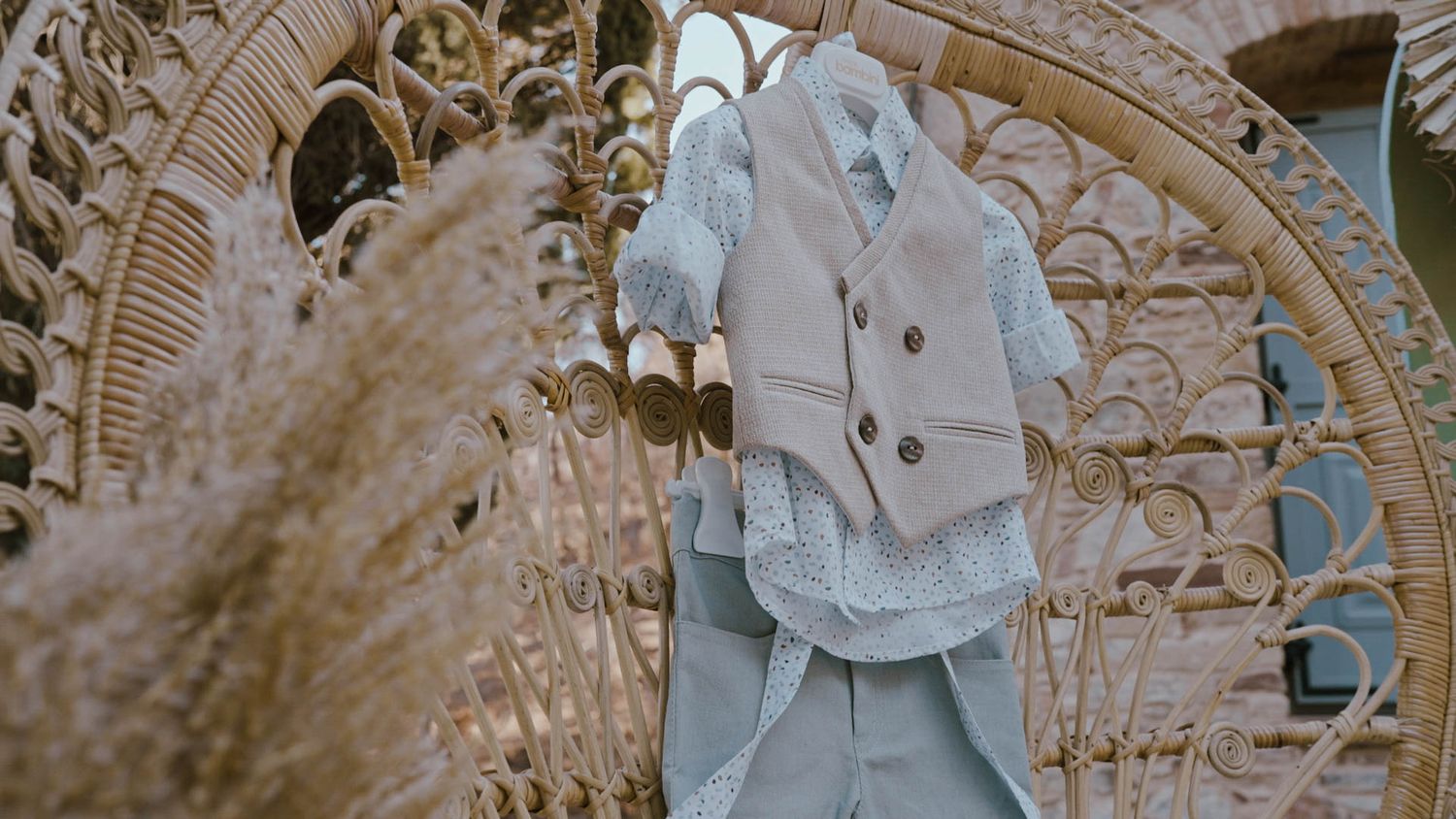 White wedding attire and shoes arranged on an ornate wicker chair with dried pampas grass decor in the background.