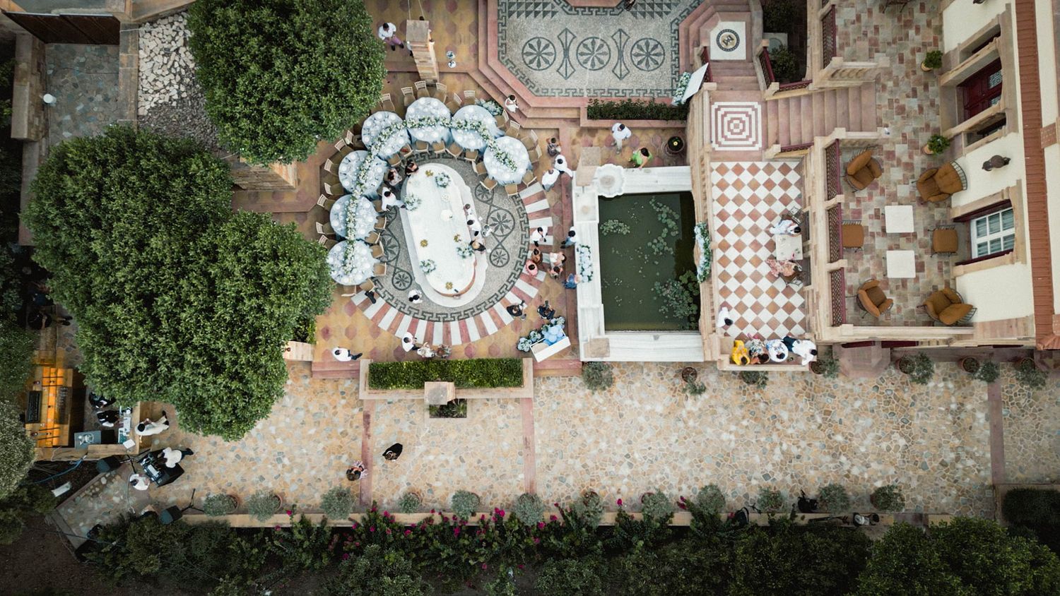 Aerial view of an outdoor dining area with circular and rectangular tables arranged on a stone patio next to greenery.