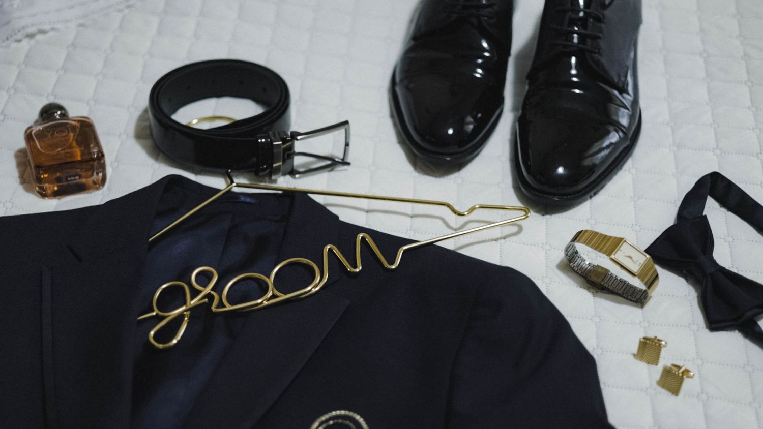 Stylish men's formal accessories including black patent leather shoes, belt, and navy blazer with groom text arranged on white floor.