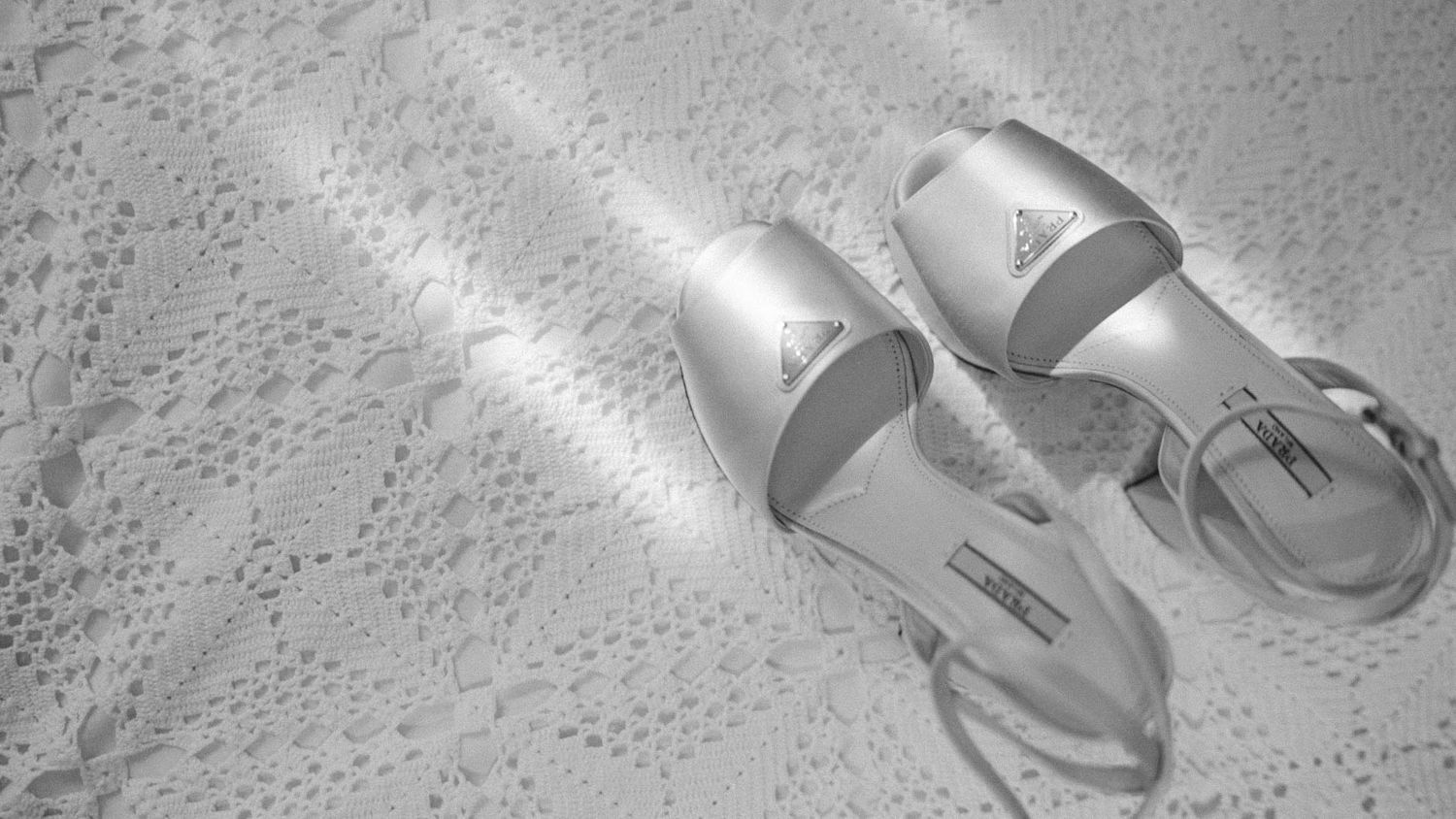 A pair of elegant silver sandals with ankle straps rests elegantly on white lace fabric in black and white tones.