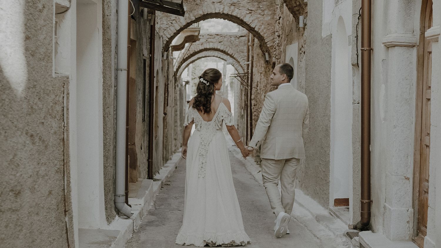Couple walking through arched stone passageway in wedding attire.