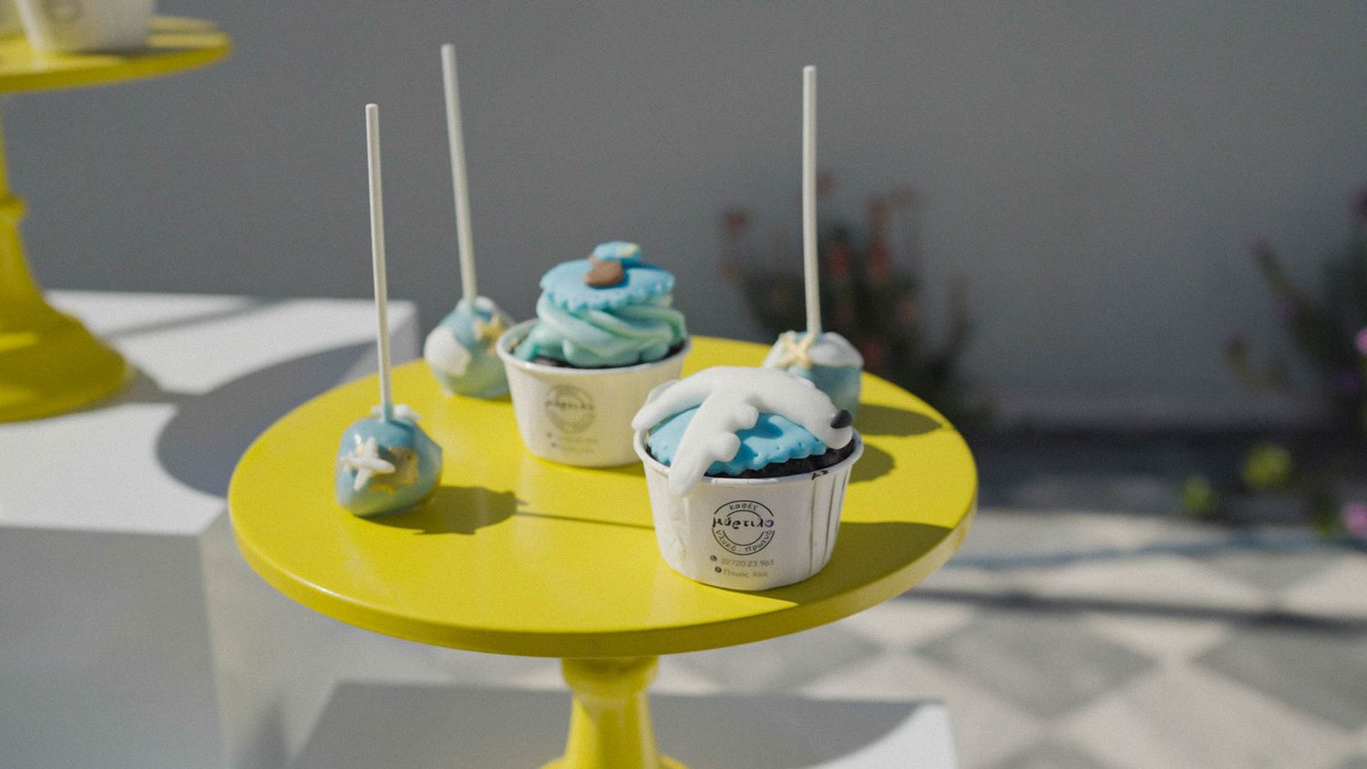 Decorated blue and white cake pops displayed on a modern yellow pedestal table in natural lighting.