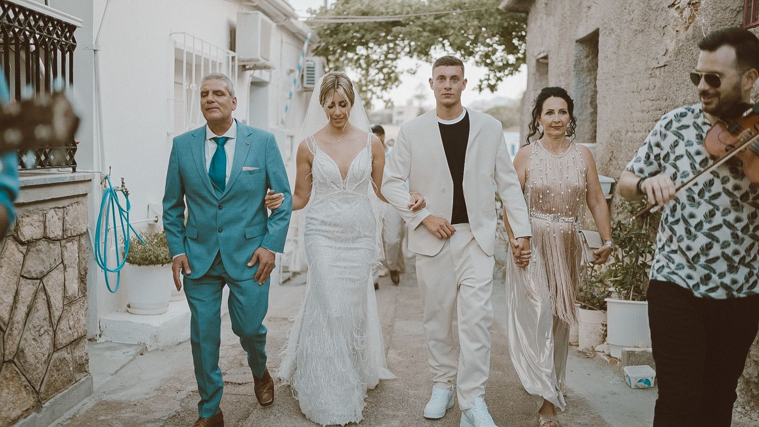 A small wedding party walks together down a Mediterranean-style street with one wearing turquoise and others in white and light tones.
