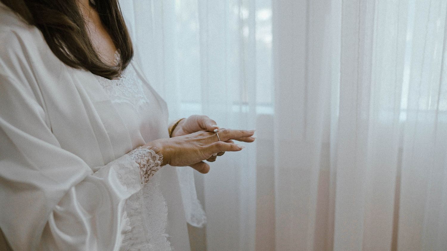 Person in white long sleeve dress holding mobile phone by sheer white curtained window during daytime.