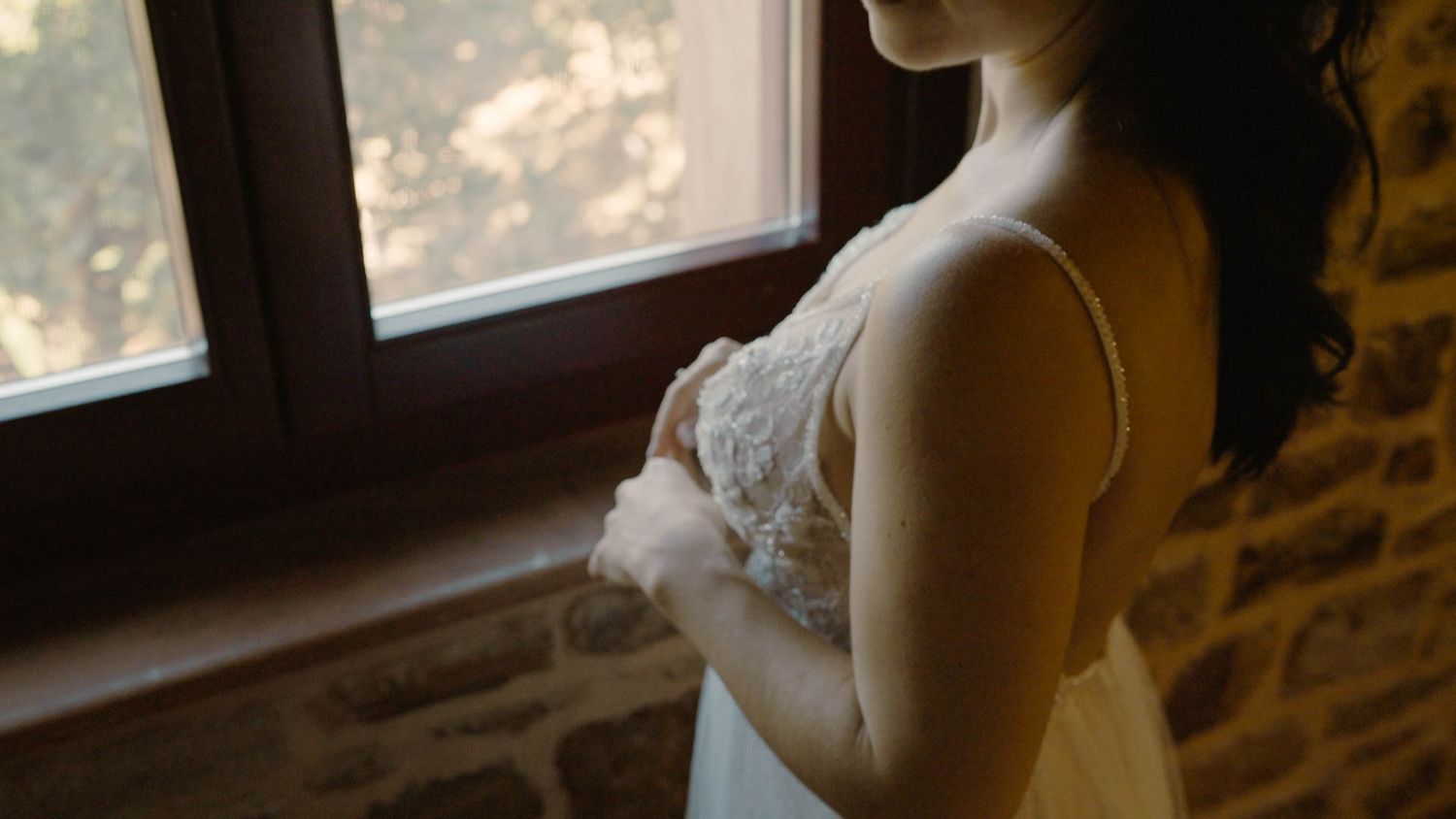 A pregnant woman in a white lace dress stands by a window in a rustic stone room, photographed in soft natural light.