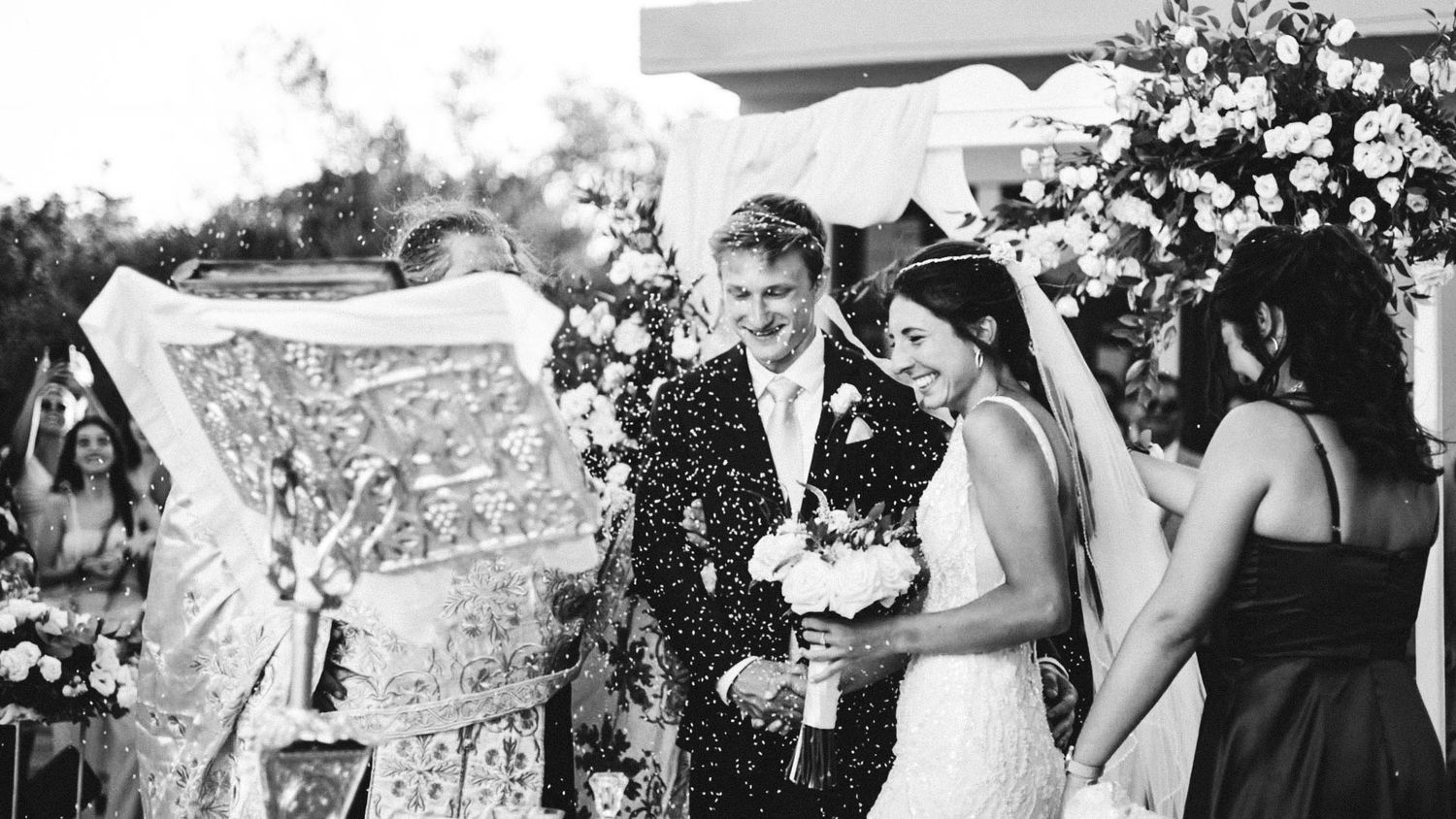 Black and white photo of newlyweds surrounded by guests throwing confetti during outdoor wedding celebration.