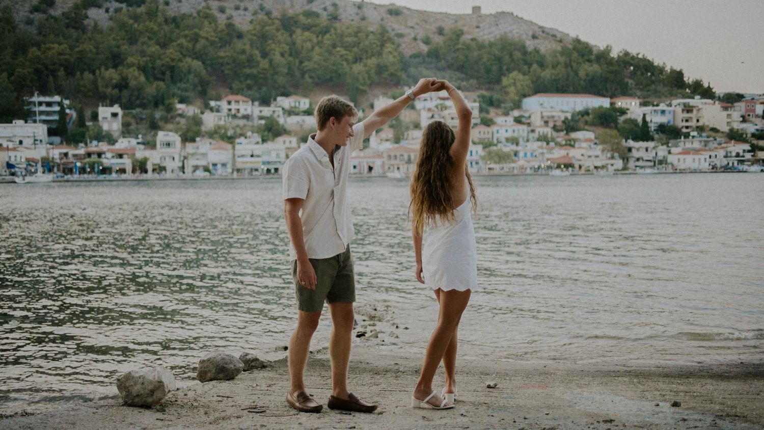 A couple dancing together on a pebbled beach at sunset with coastal mountain and town views in the background.