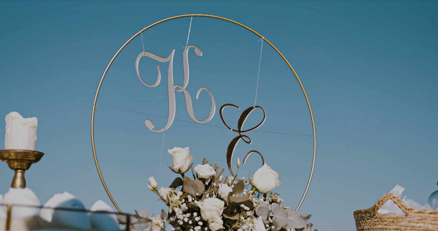 Gold circular monogram with initials K and E against a blue sky backdrop with white floral decorations.
