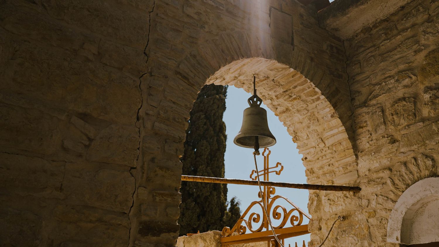 A vintage bell hangs from a stone archway against a blue sky, featuring ornate wrought iron decorative details.